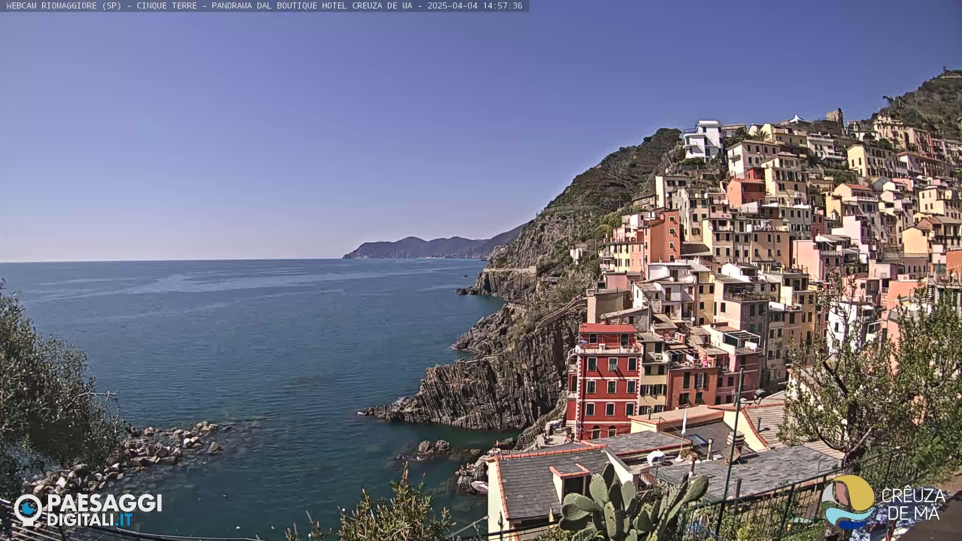 A colorful hillside village clings to a rocky coast overlooking a calm, blue sea under a clear, sunny sky.