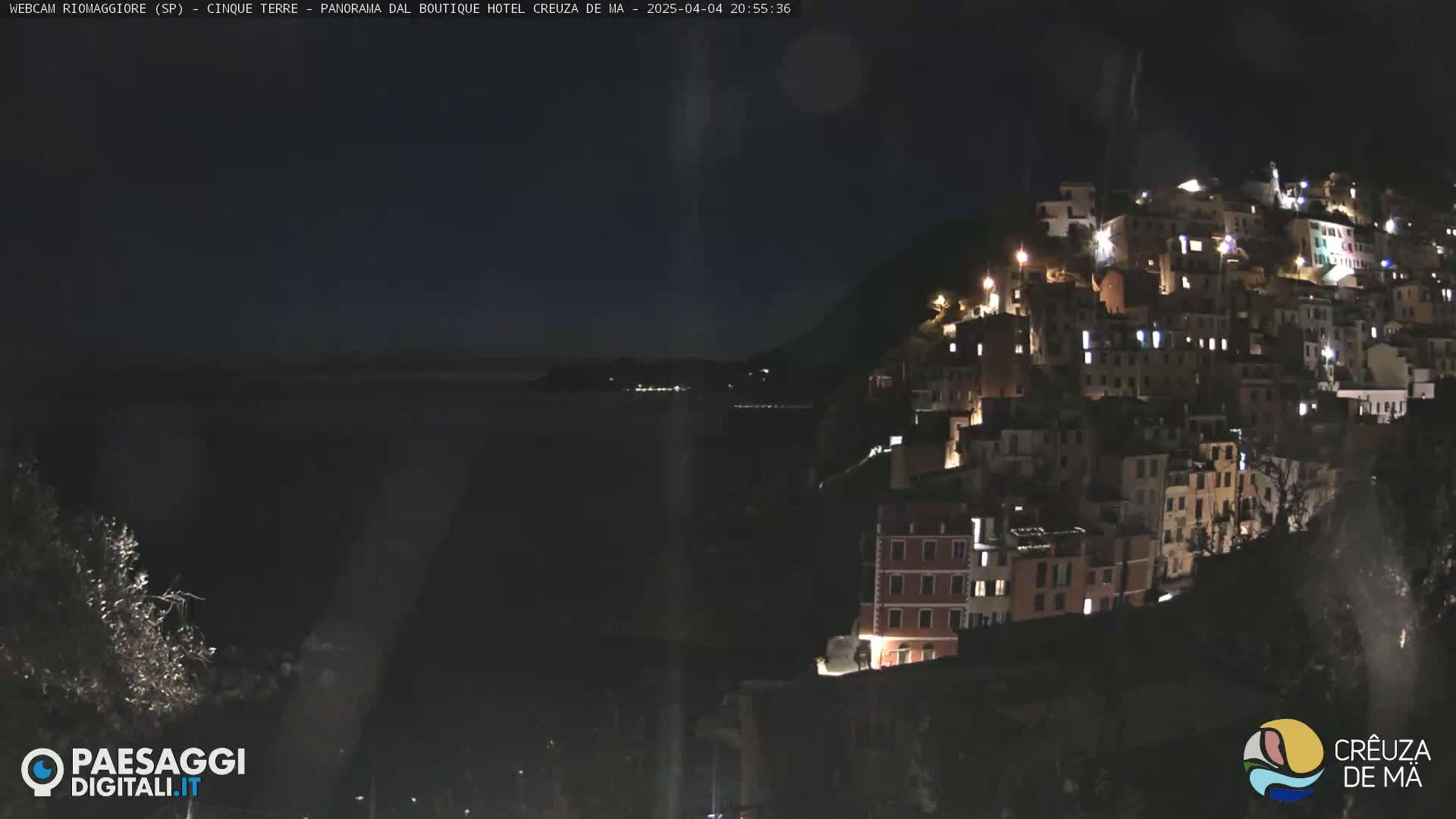 A nighttime view of a hilltop village illuminated by lights, overlooking a dark body of water under a clear, dark sky.
