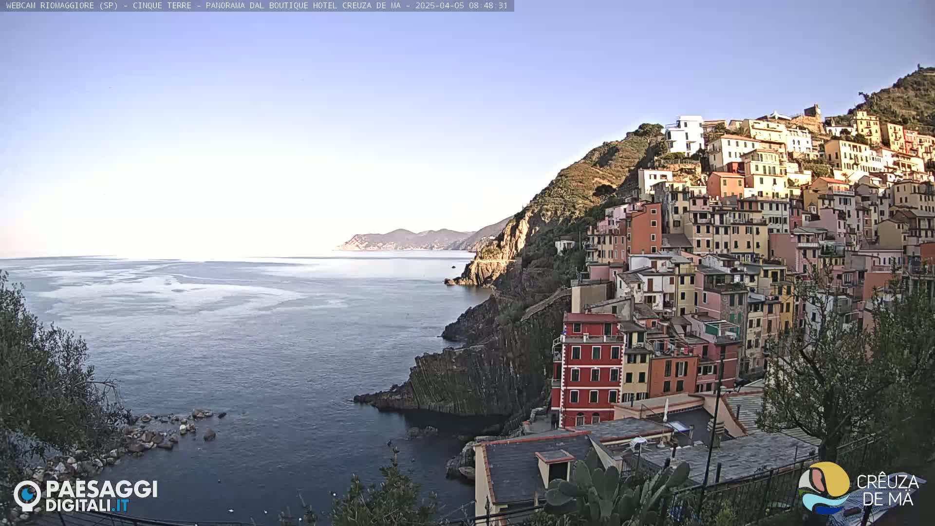 A colorful village clings to a steep hillside overlooking a calm sea under a clear, bright sky.