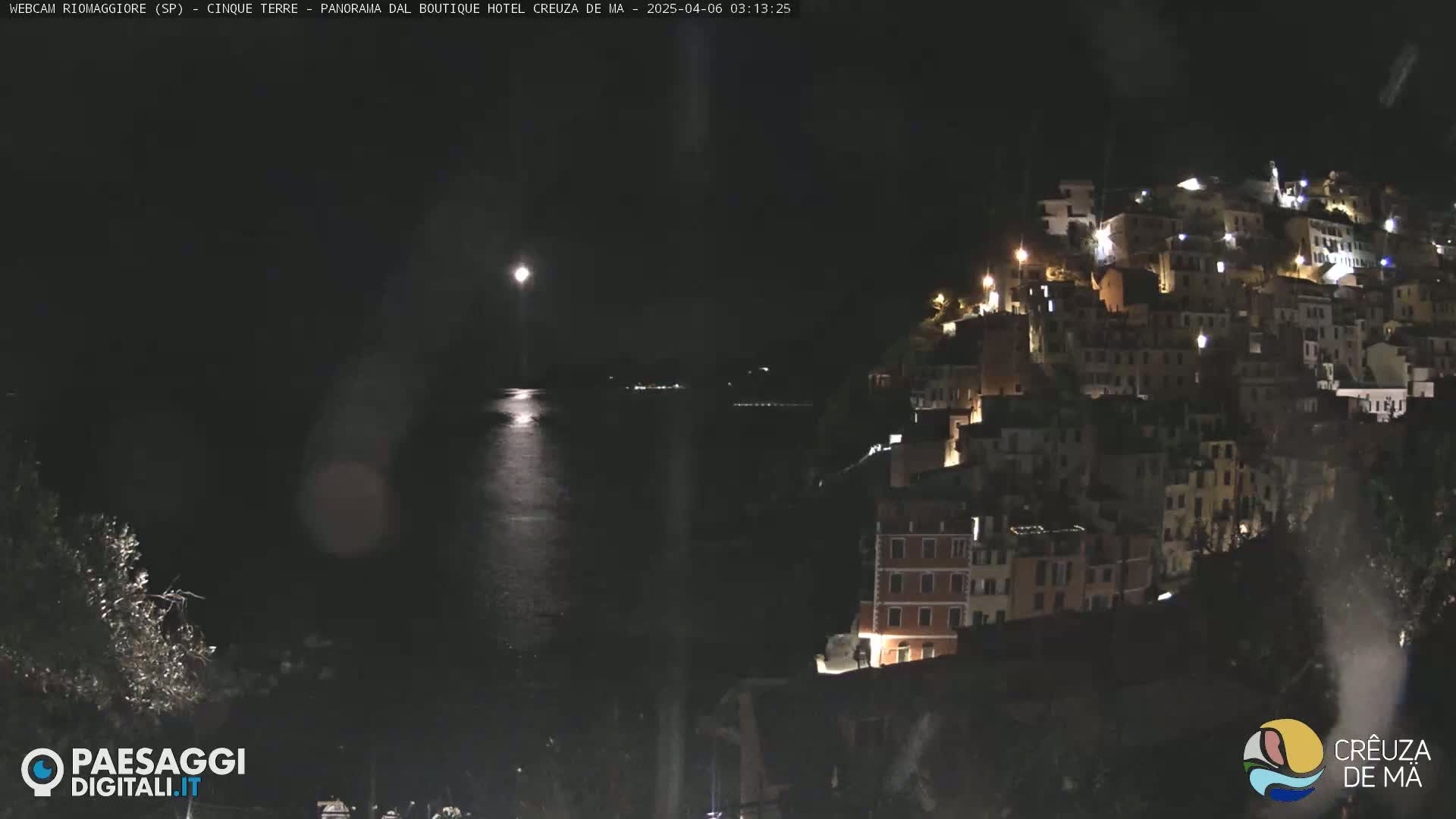 A nighttime view of a hillside town illuminated by lights, overlooking a calm body of water under a bright moon, with light rain or snow falling.