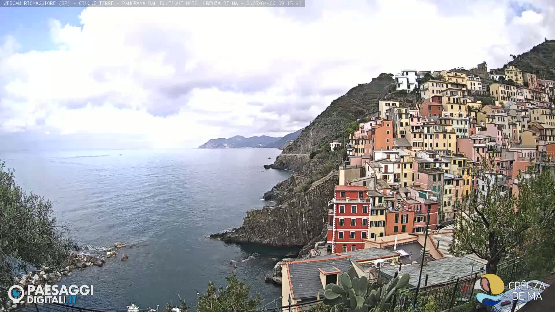 A coastal village of colorful buildings climbs a steep hillside overlooking a calm sea under a partly cloudy sky.