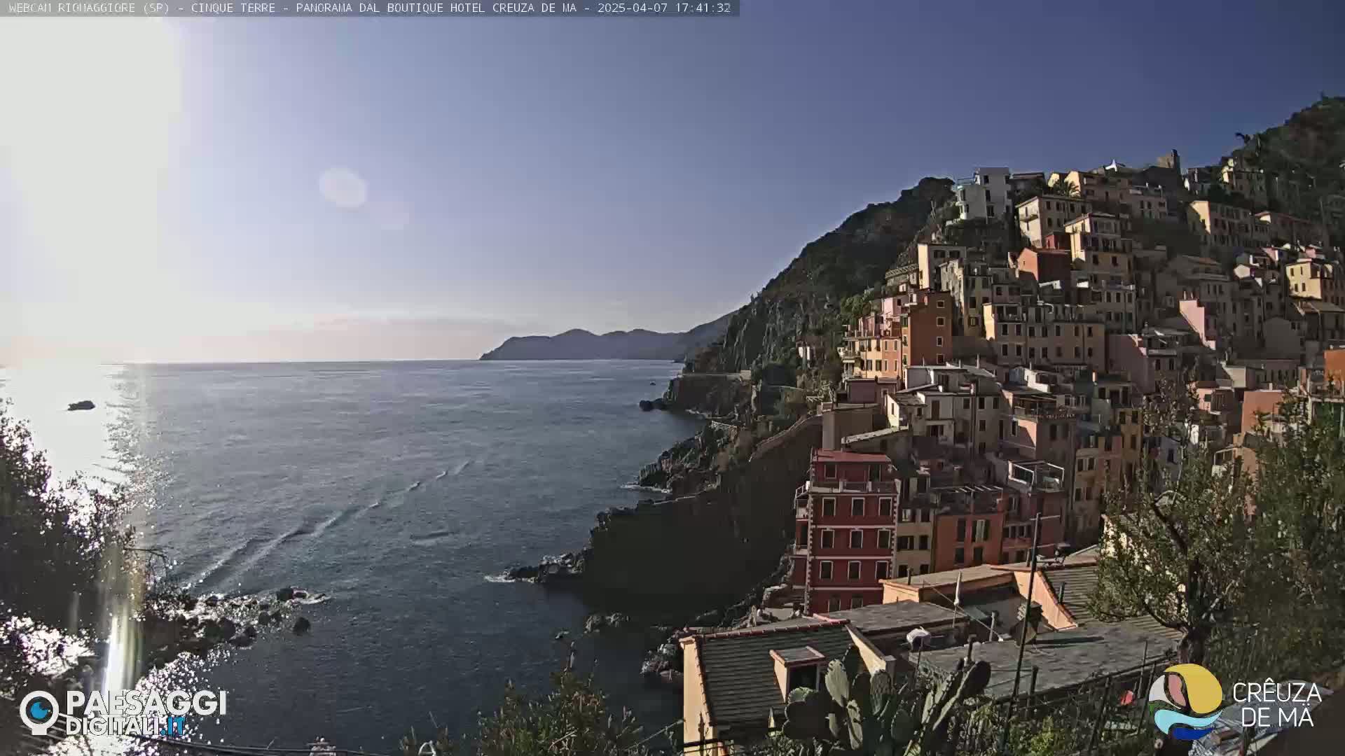 A colorful hillside town overlooks a calm, blue sea under a clear, sunny sky.