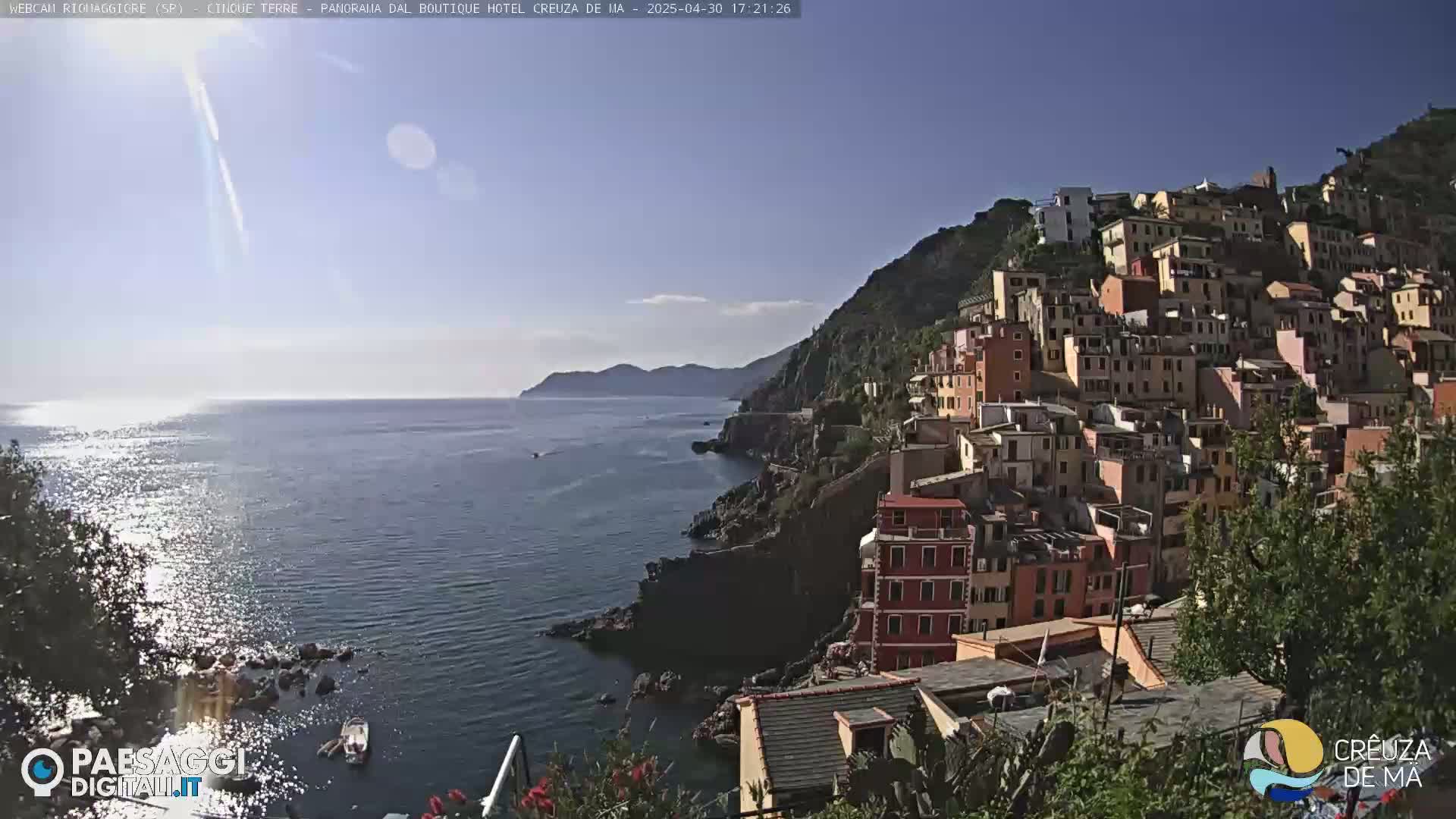 A sunlit coastal town of colorful buildings clings to a hillside overlooking a calm sea under a clear blue sky.