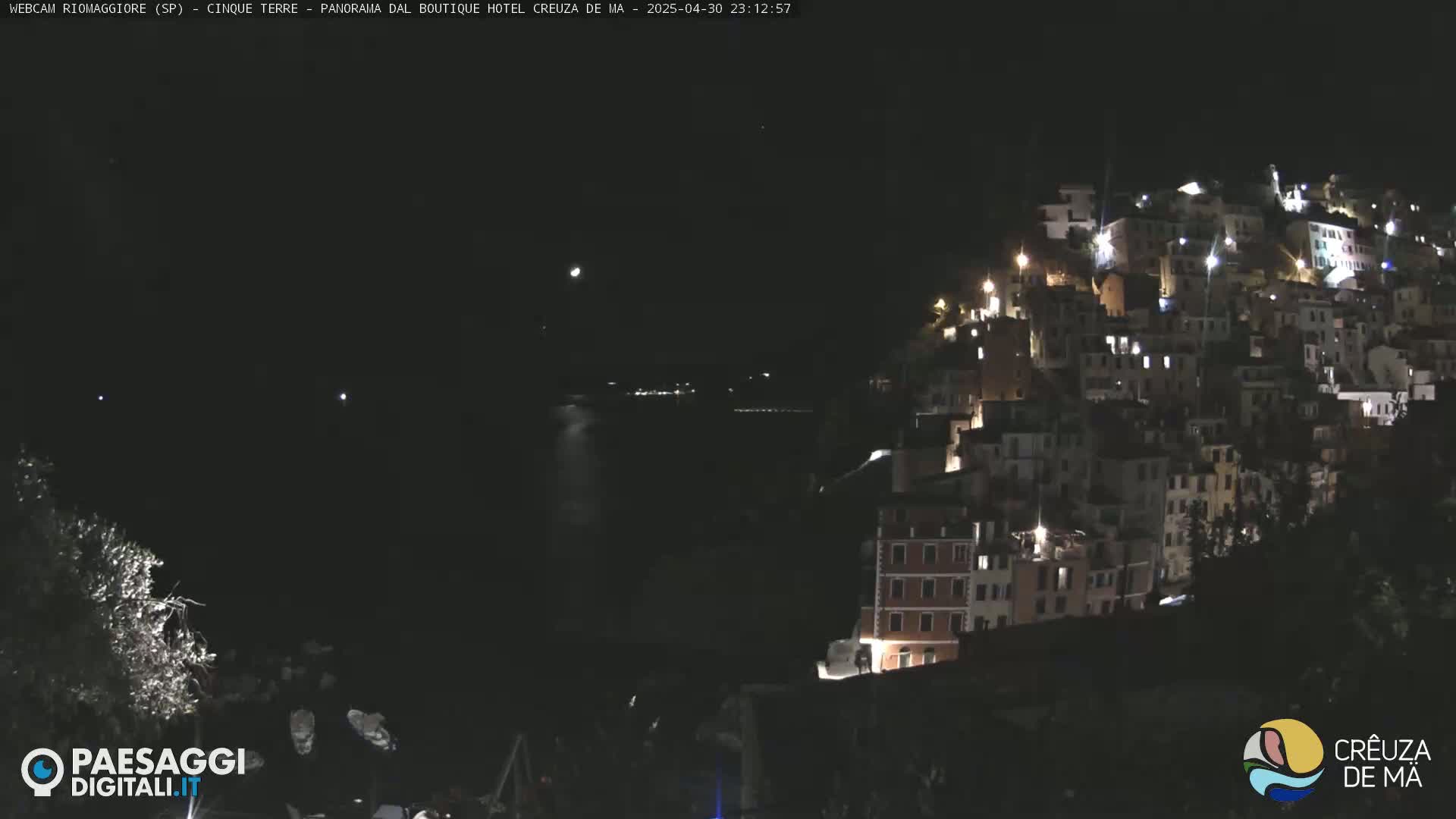 A nighttime view of a hillside town illuminated by lights, with a dark body of water visible in the distance under a clear, dark sky.
