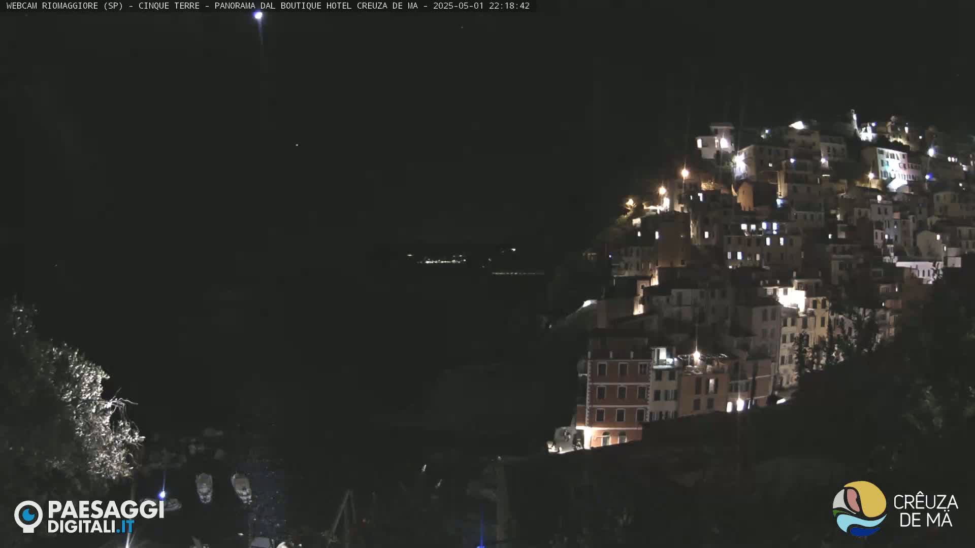 A nighttime view of a hillside village illuminated by lights, overlooking a dark body of water under a clear, dark sky.