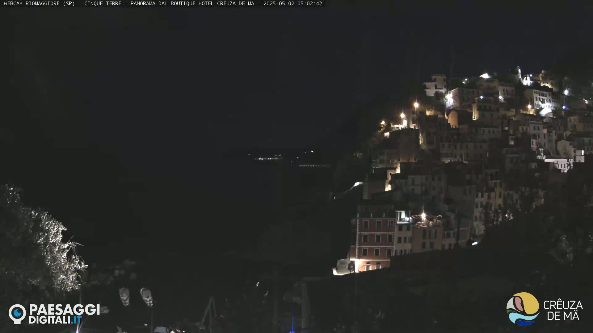 A nighttime view of a hillside town illuminated by lights, overlooking a dark body of water under a clear, dark sky.
