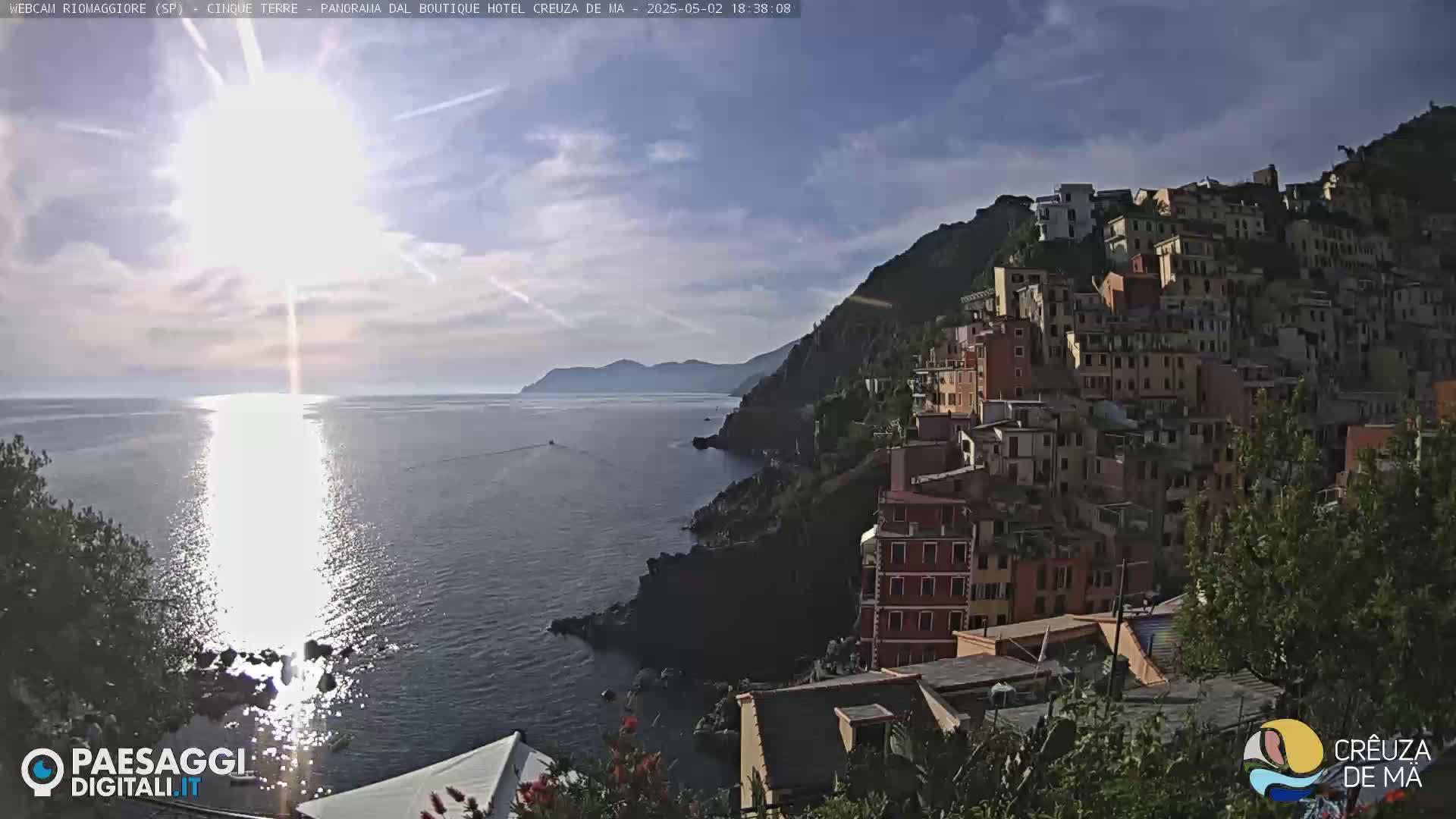 A sunlit seaside town clings to a steep hillside overlooking a calm, sparkling sea under a partly cloudy sky.