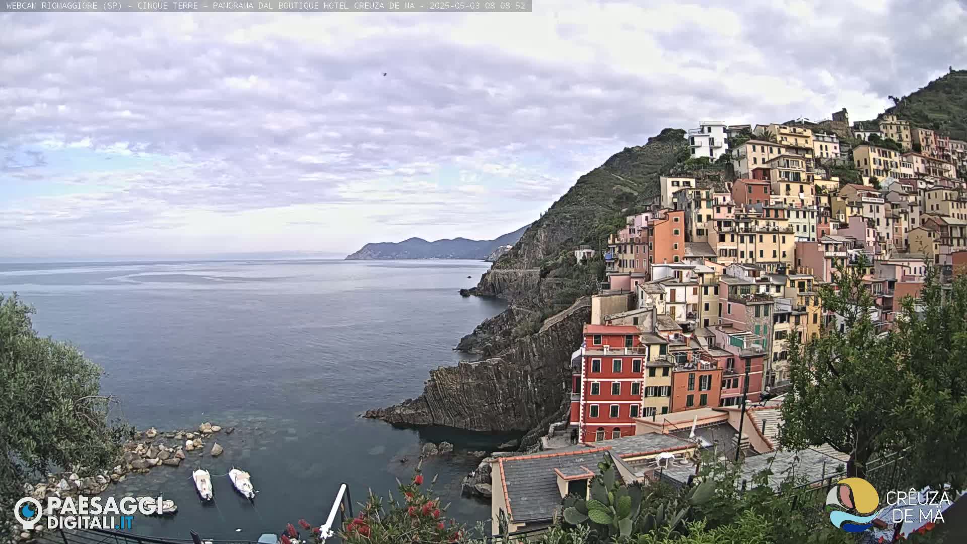 A coastal town with colorful buildings clustered on a hillside overlooking a calm sea under a mostly cloudy sky.