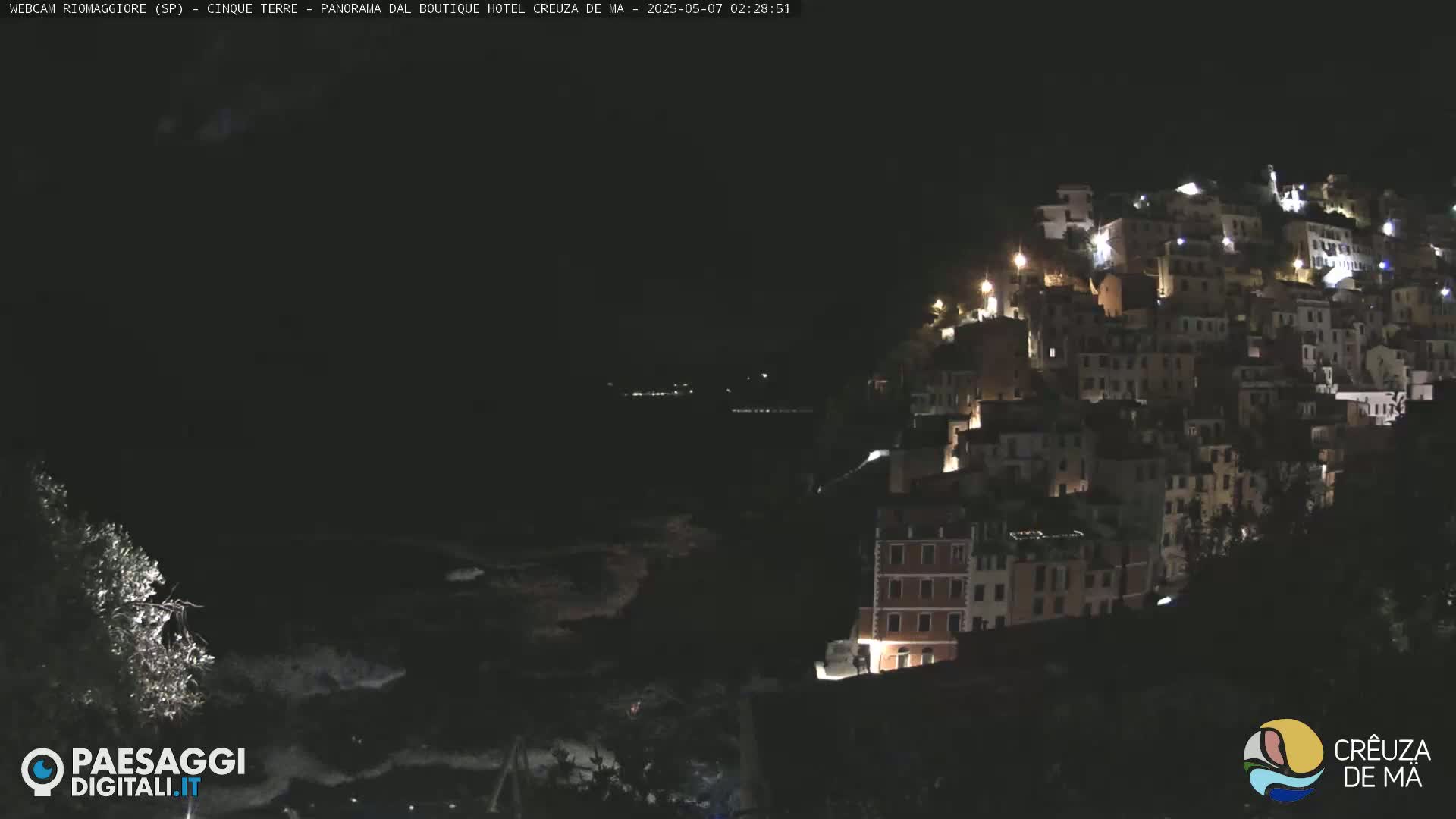 A nighttime view of a hillside village with many lit windows, overlooking a dark body of water.