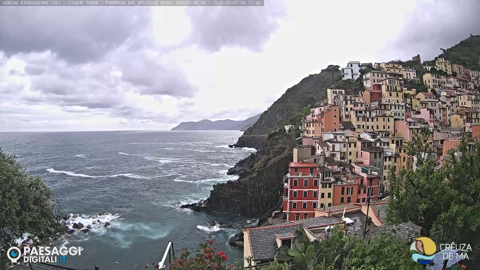 A coastal village of colorful buildings clings to a steep hillside overlooking a dark blue, choppy sea under a cloudy sky.