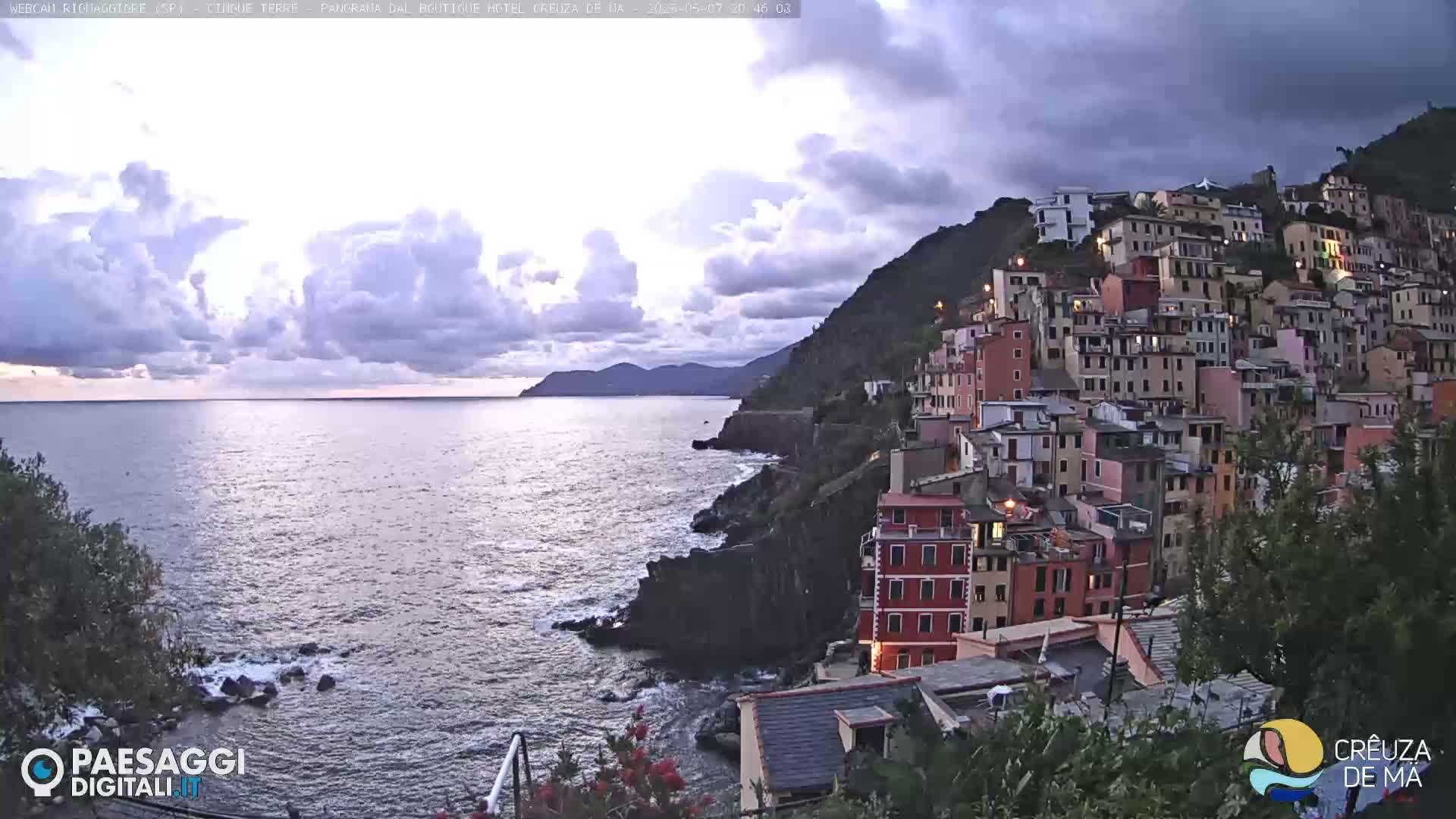 A coastal village of colorful buildings clings to a hillside overlooking a calm sea under a partly cloudy sky.