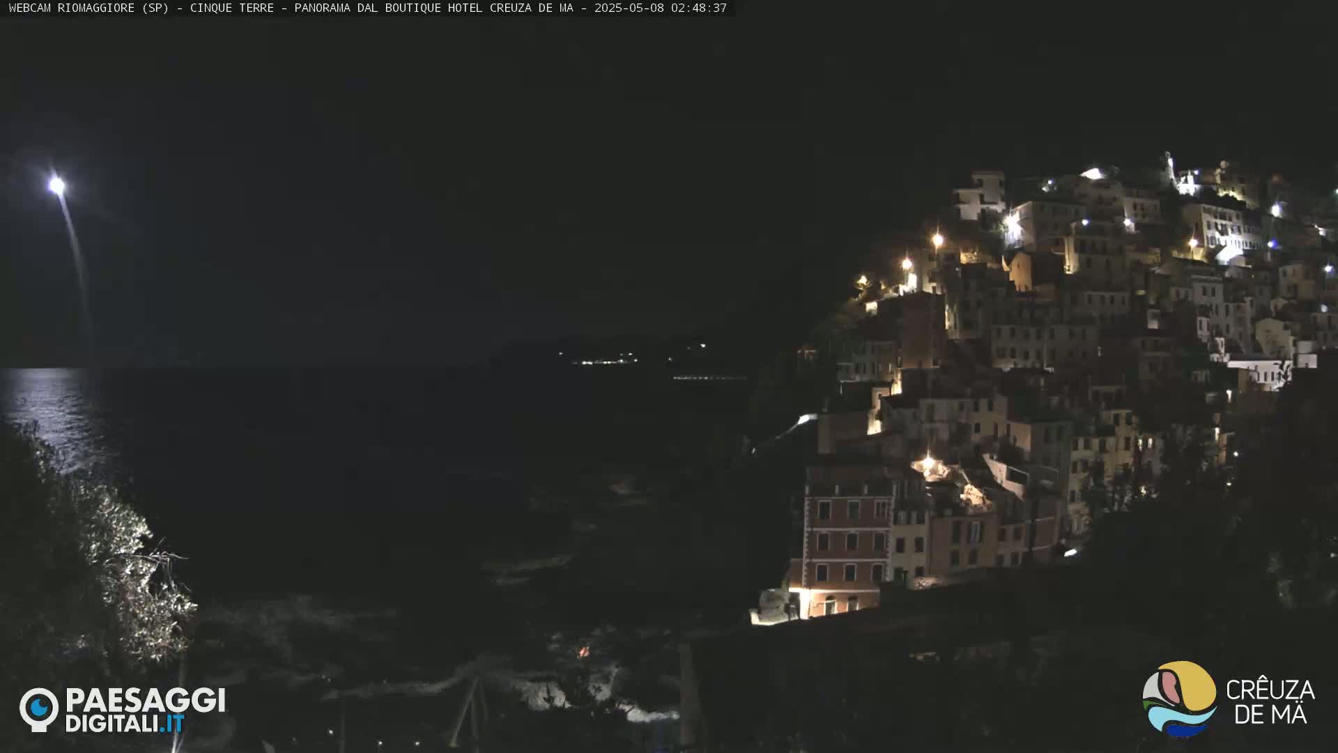 A brightly lit town on a hill overlooking a dark, calm ocean at night, with a bright light visible in the distance over the water.