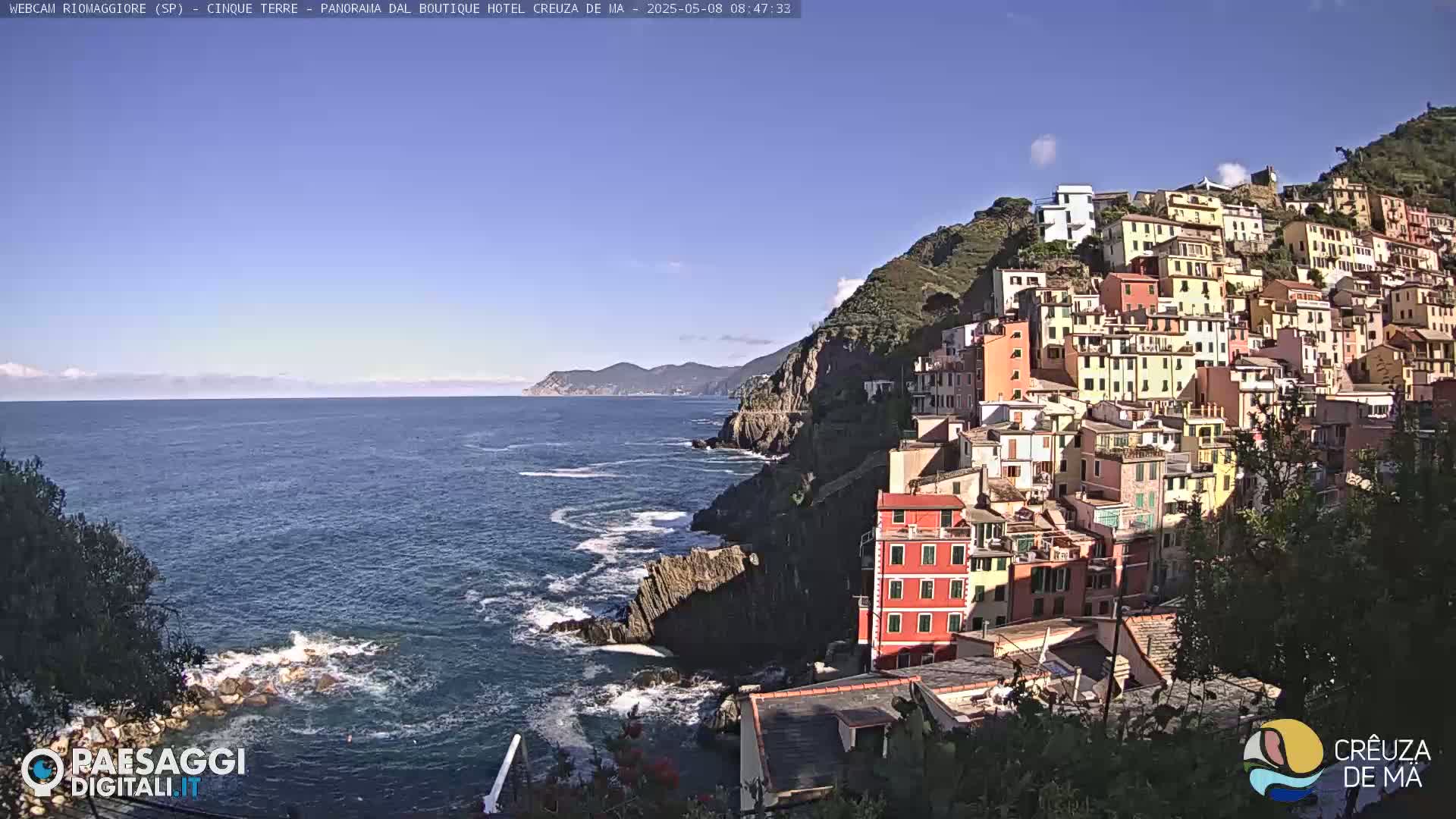 A colorful hillside village overlooks a calm, blue sea under a clear, sunny sky.