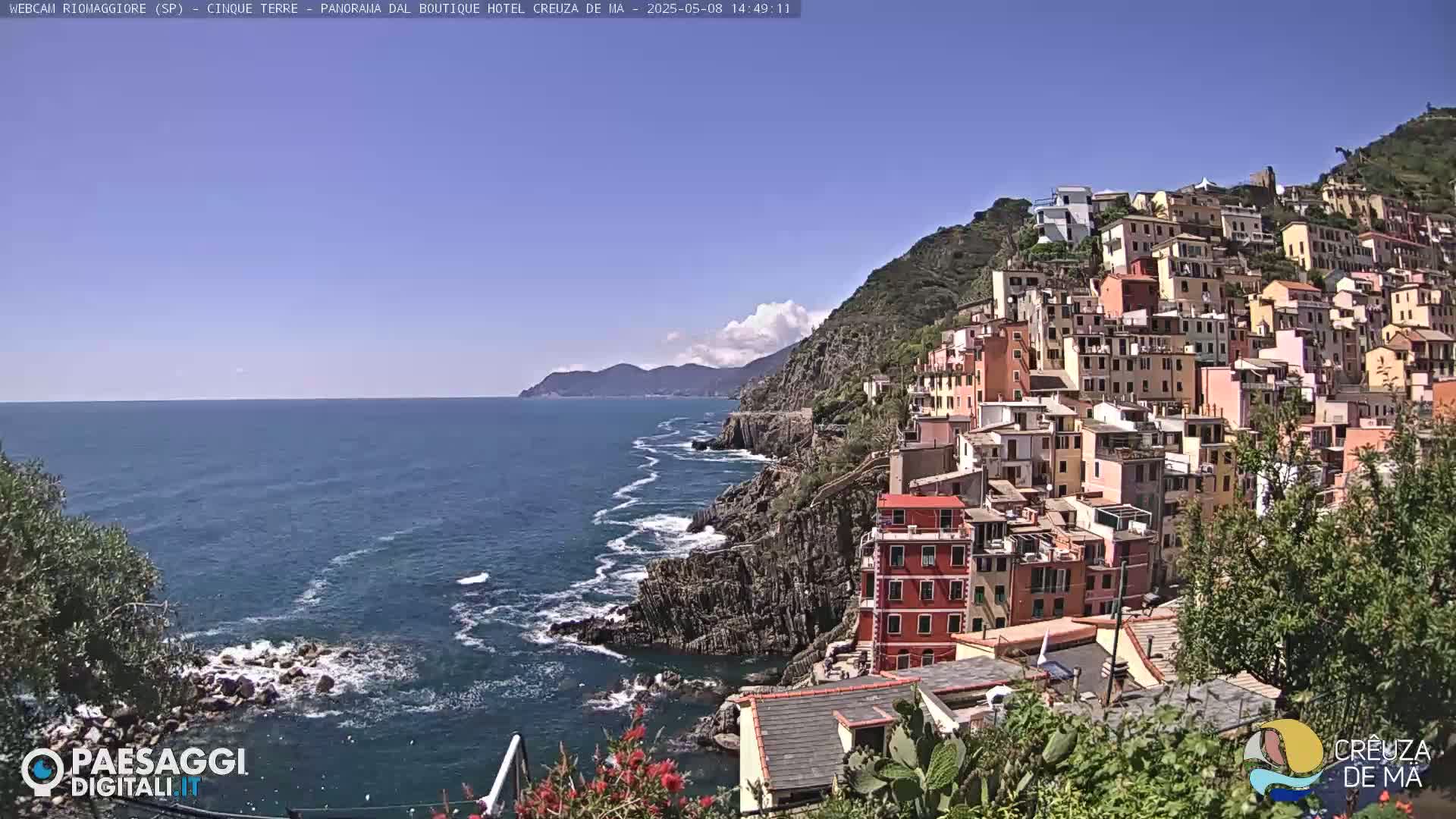 A colorful village clings to a steep, rocky hillside overlooking a blue sea with some small waves under a mostly clear sky.