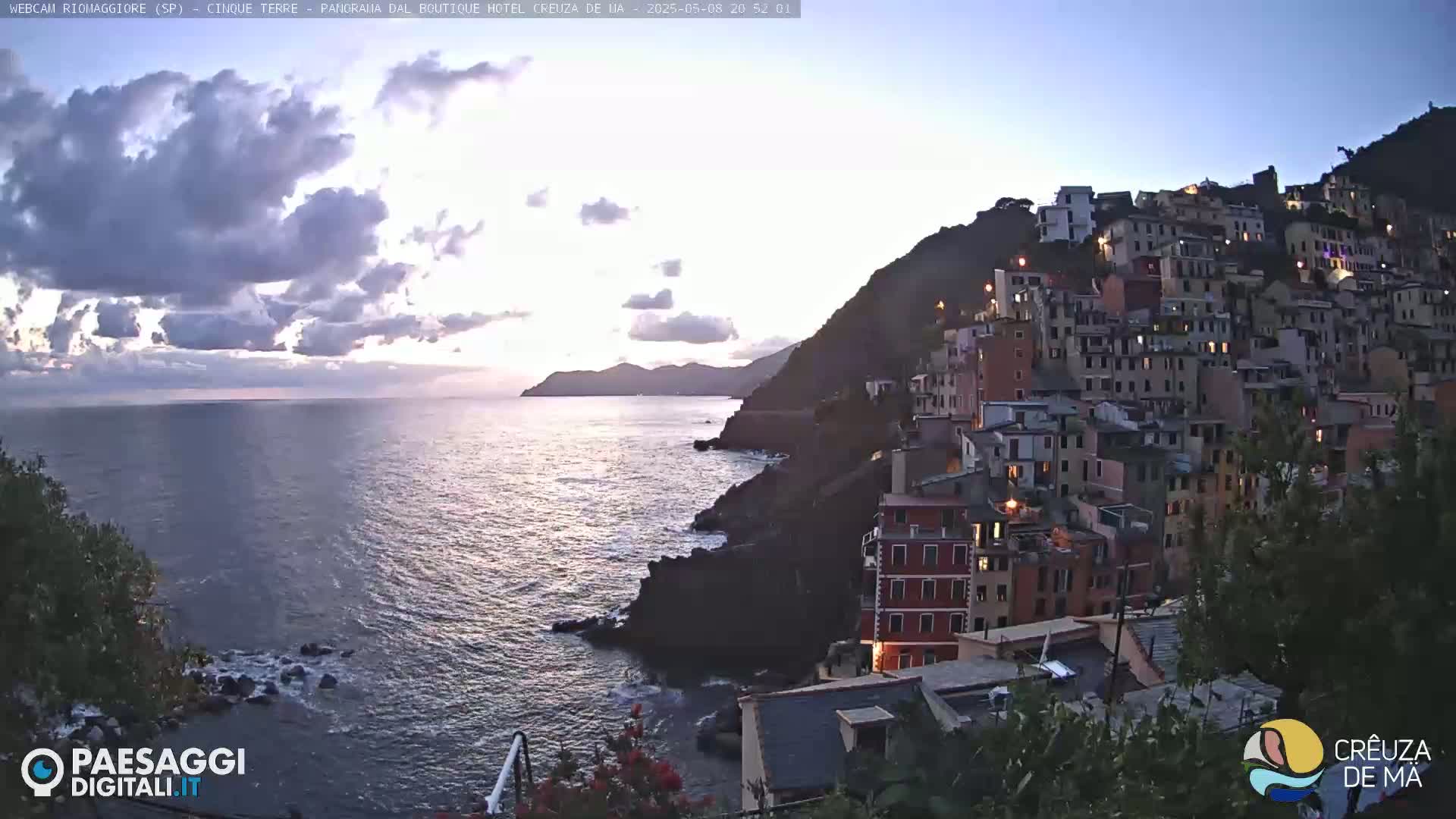 A coastal town is nestled on a hillside overlooking a calm sea under a partly cloudy twilight sky.