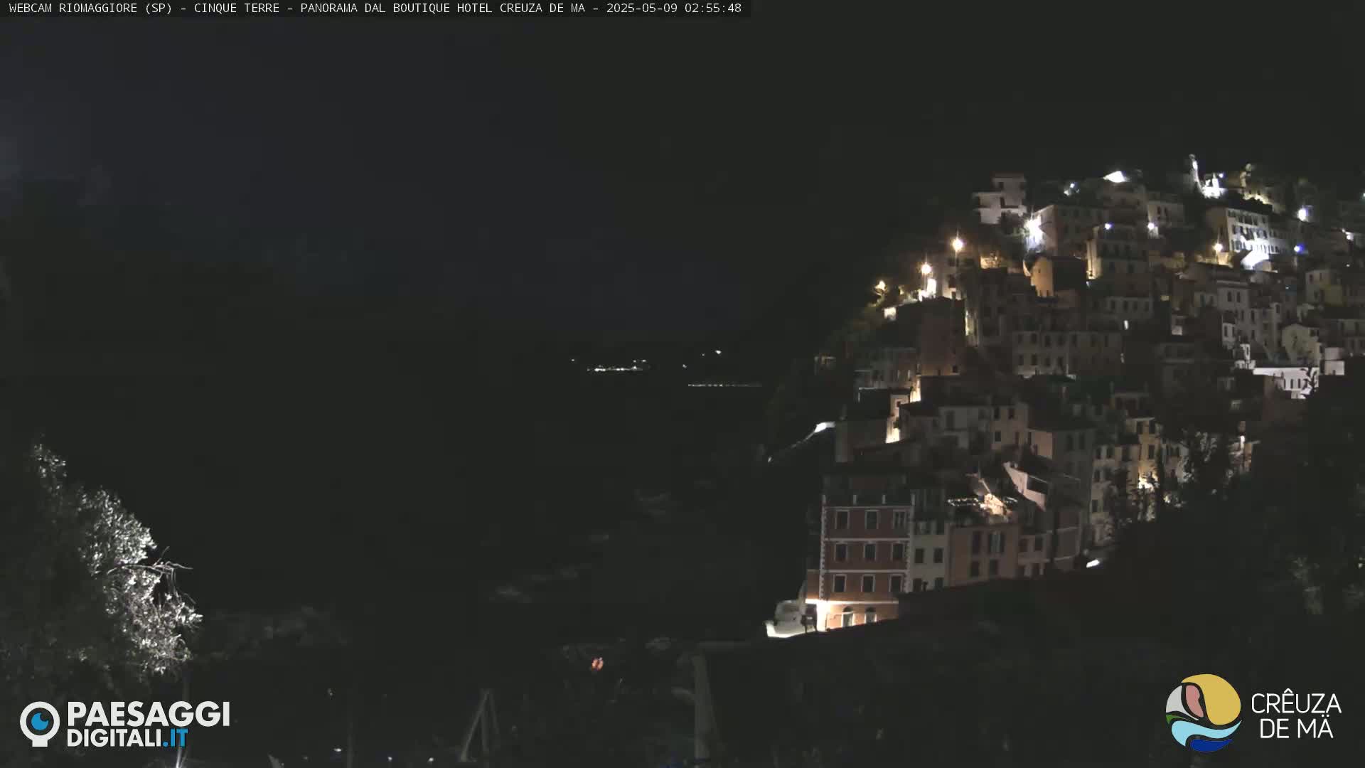 A hillside village is illuminated at night, overlooking a dark body of water under a clear, dark sky.