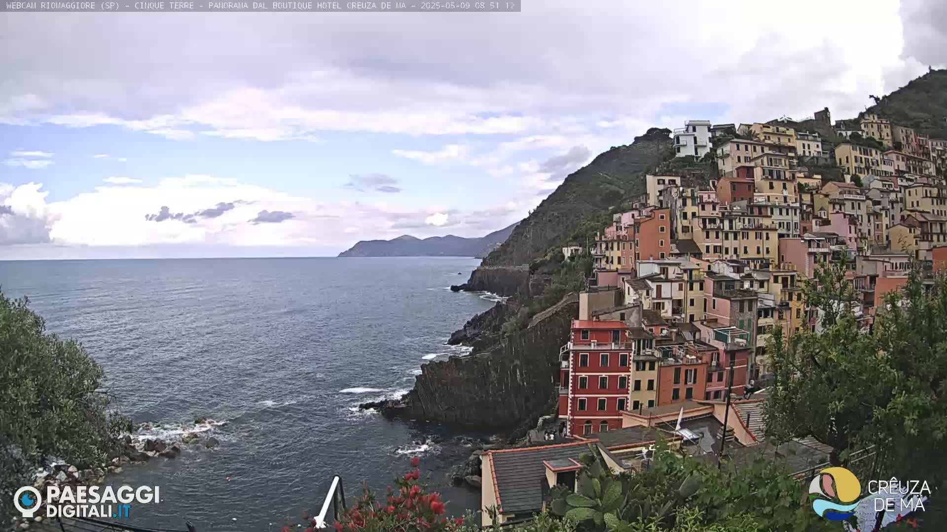 A coastal village of colorful buildings clings to a steep hillside overlooking a calm sea under a partly cloudy sky.