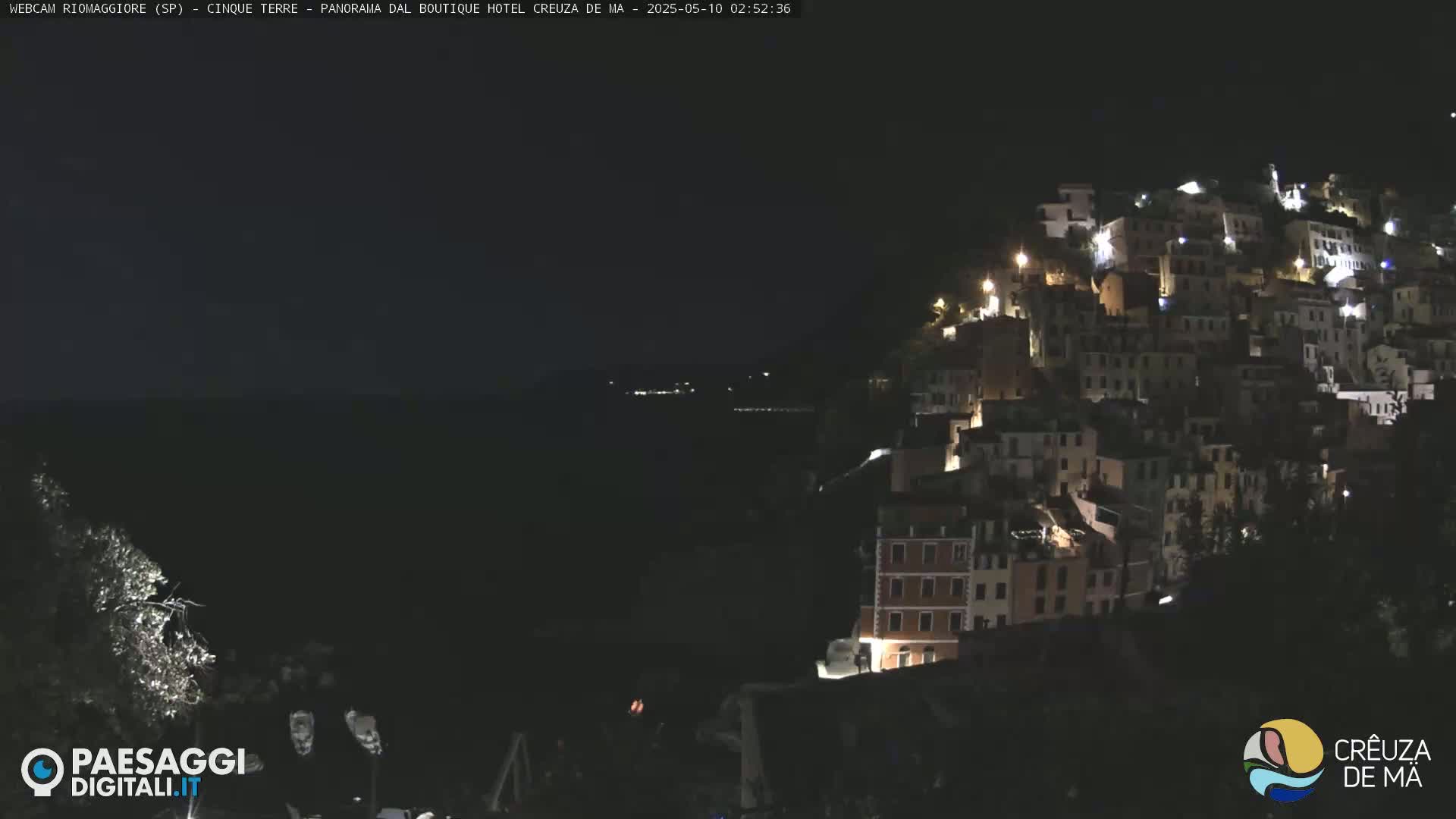 A nighttime view of a hillside town illuminated by lights, overlooking a dark body of water under a clear sky.