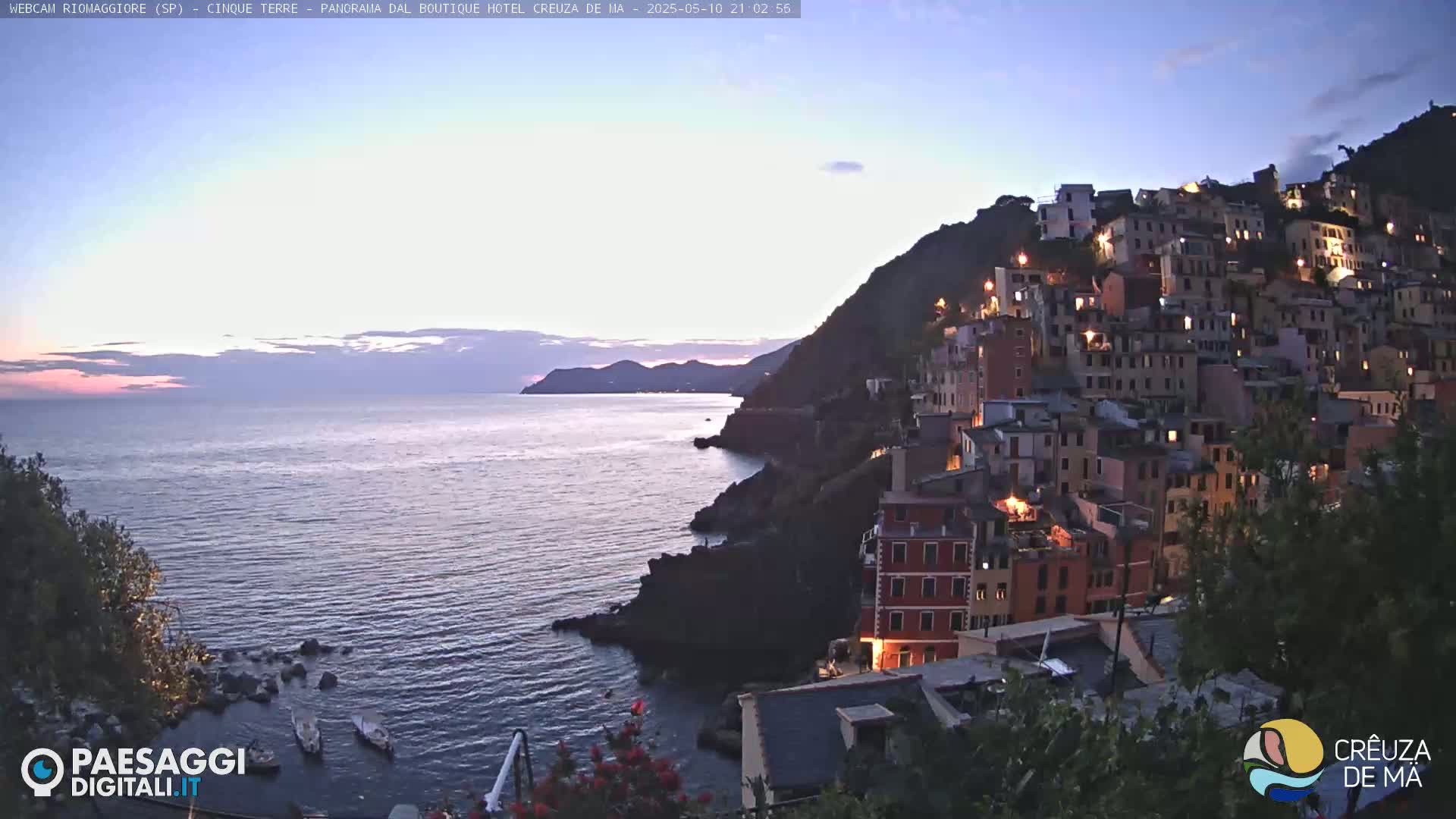 A twilight view shows a hillside town's lights glowing against a dark, rocky coastline and a calm, dark-purple sea under a dusky sky.