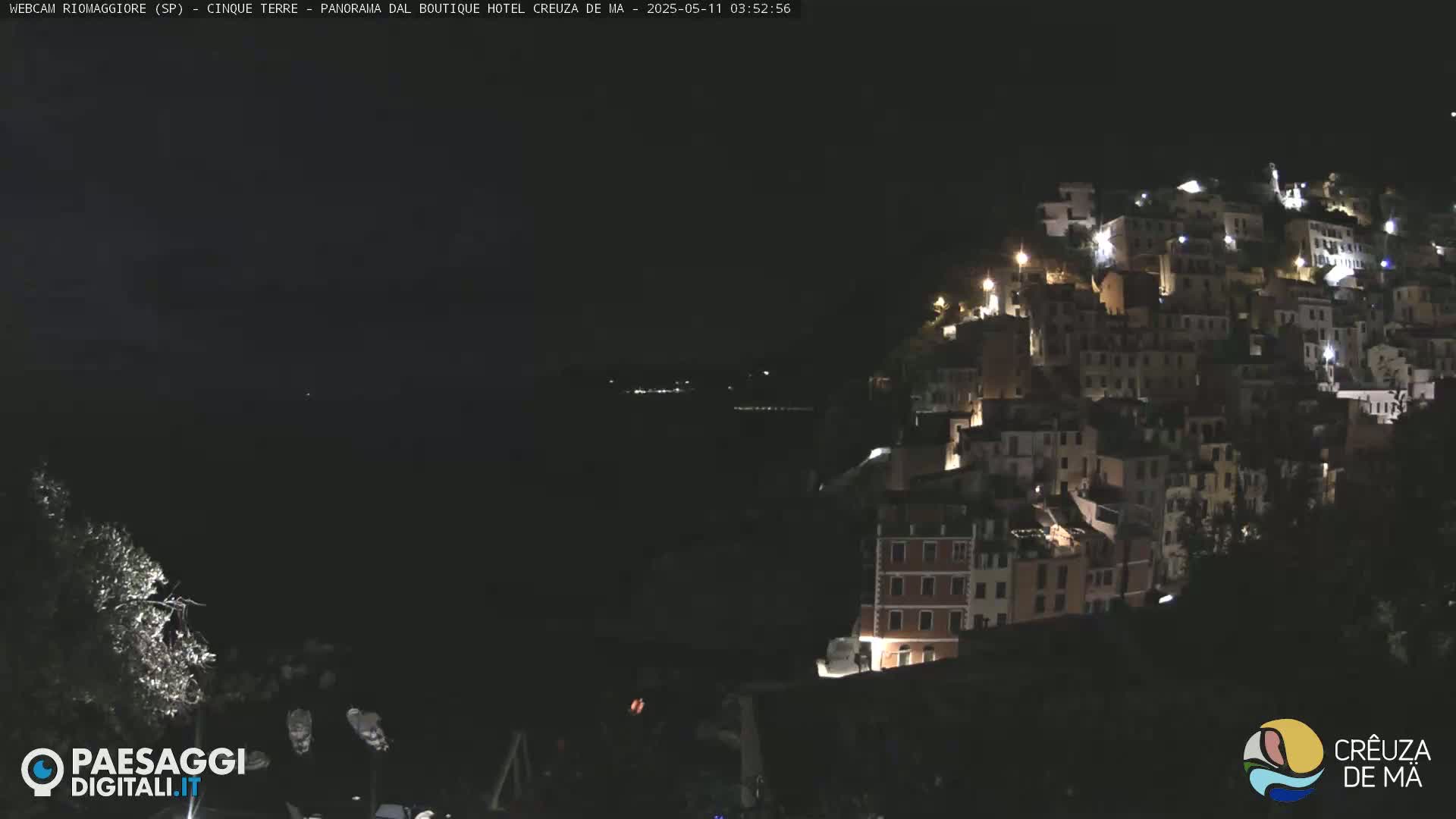 A nighttime view of a hillside town illuminated by lights, overlooking a dark body of water.