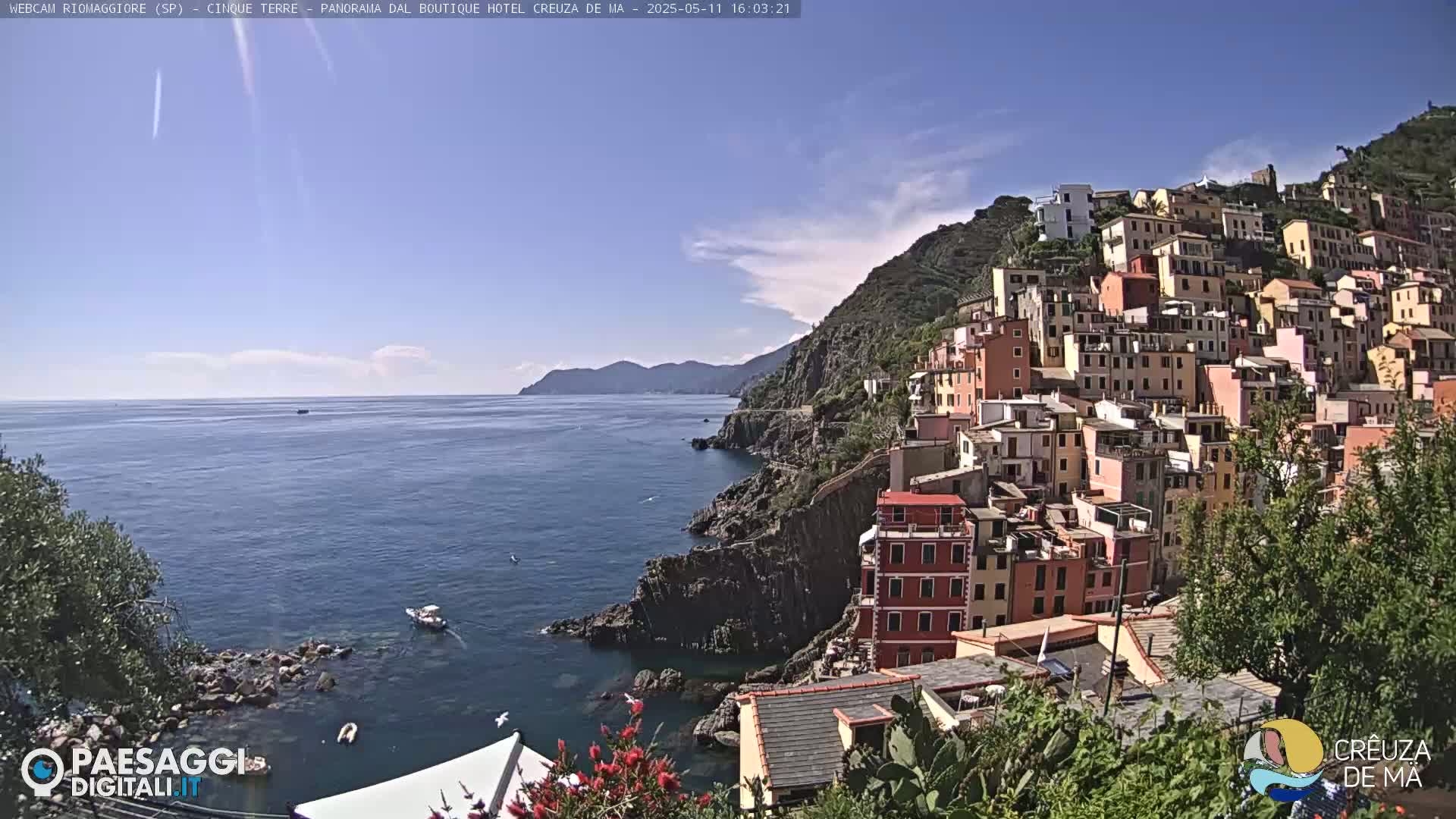 A picturesque coastal town with colorful buildings clinging to a steep hillside overlooking a calm blue sea under a mostly sunny sky.