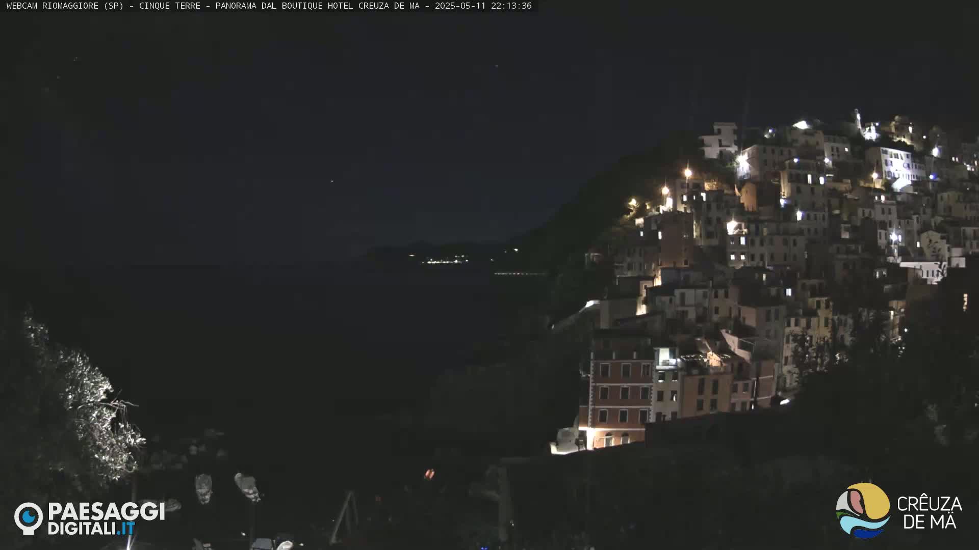 A nighttime view of a hillside town illuminated by lights, overlooking a dark body of water under a clear sky.