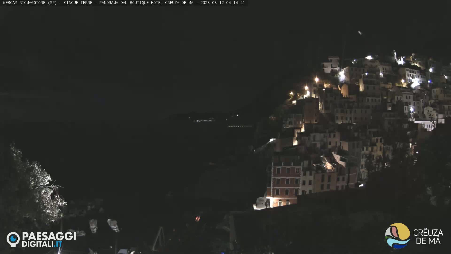A nighttime, dimly lit coastal town clings to a hillside, overlooking a dark body of water.