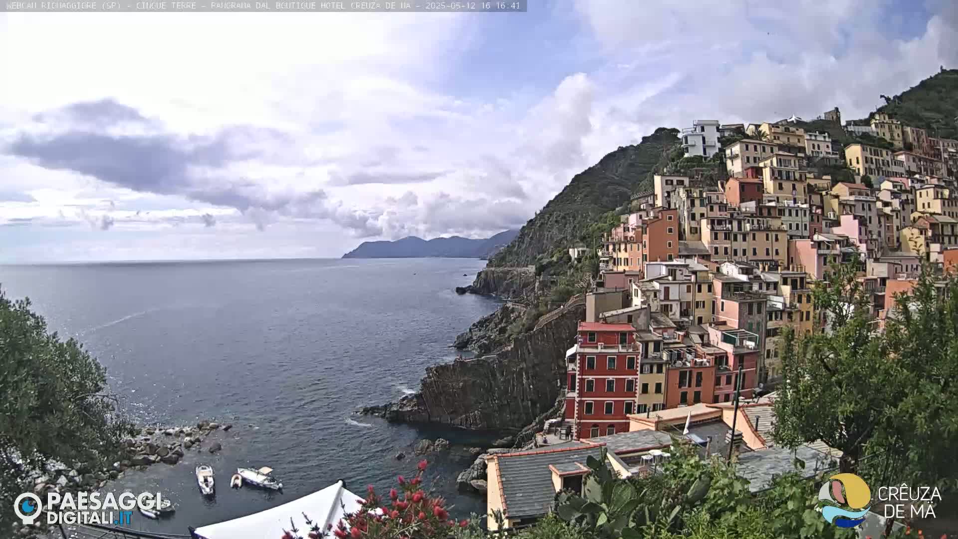 A coastal village of colorful buildings climbs a steep hillside overlooking a calm sea under a partly cloudy sky.