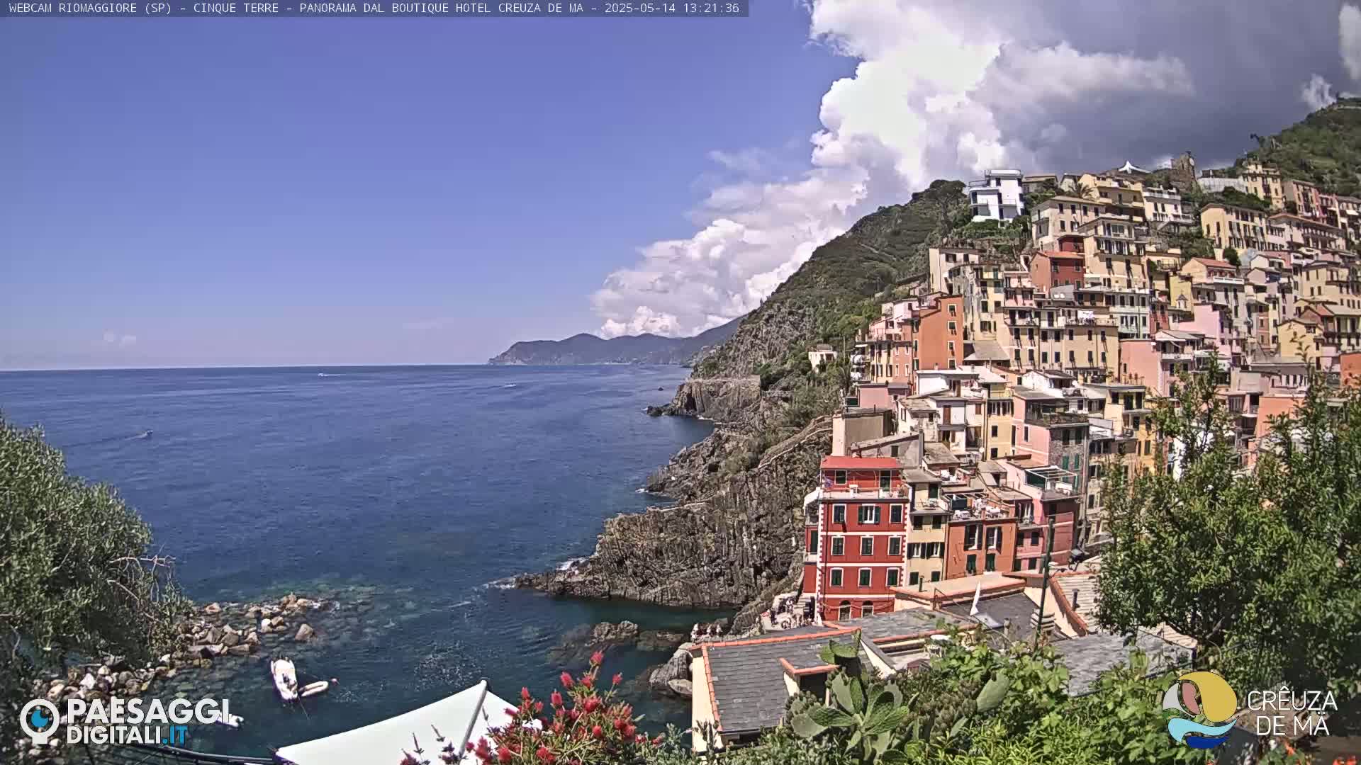 A coastal village of colorful buildings clings to a steep hillside overlooking a calm blue sea under a partly cloudy sky.