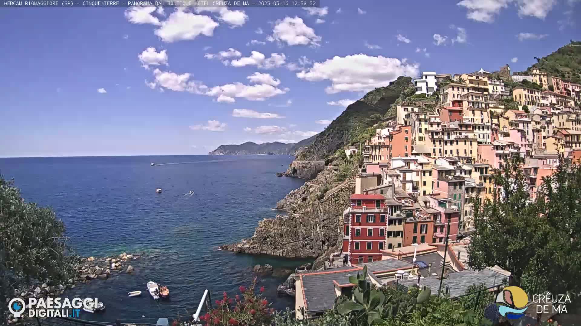 A colorful village clings to a rocky hillside overlooking a calm blue sea under a partly cloudy sky.