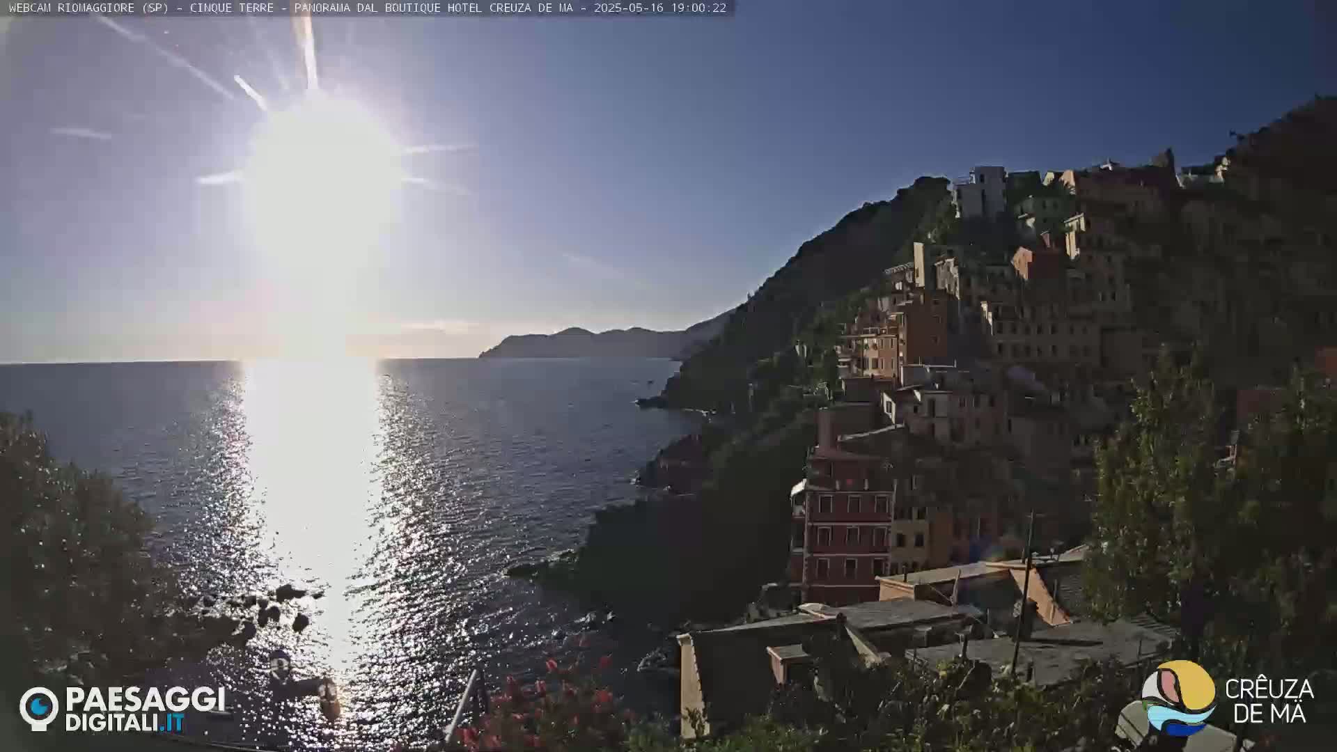 A bright, sunny day reveals a coastal village clinging to a hillside overlooking a sparkling sea with the sun reflecting on the water.