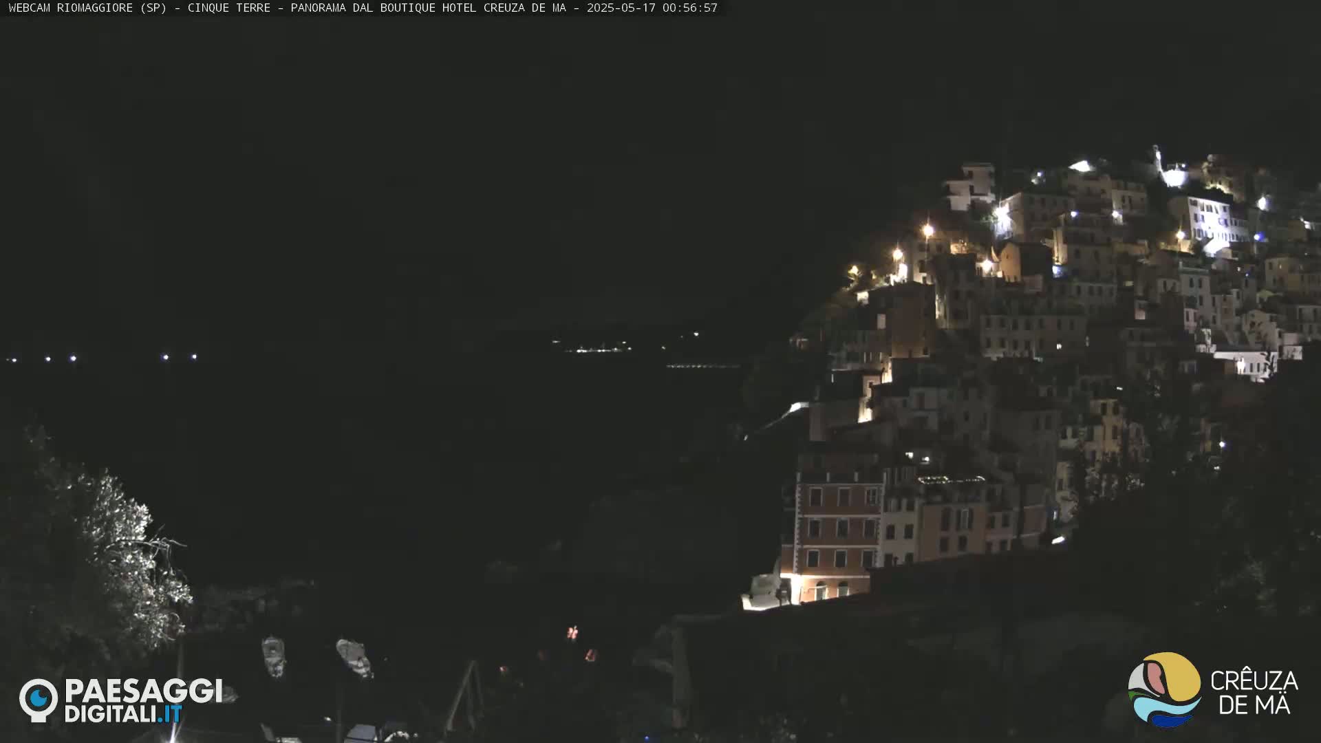 A nighttime view of a hillside town illuminated by lights, overlooking a dark body of water.