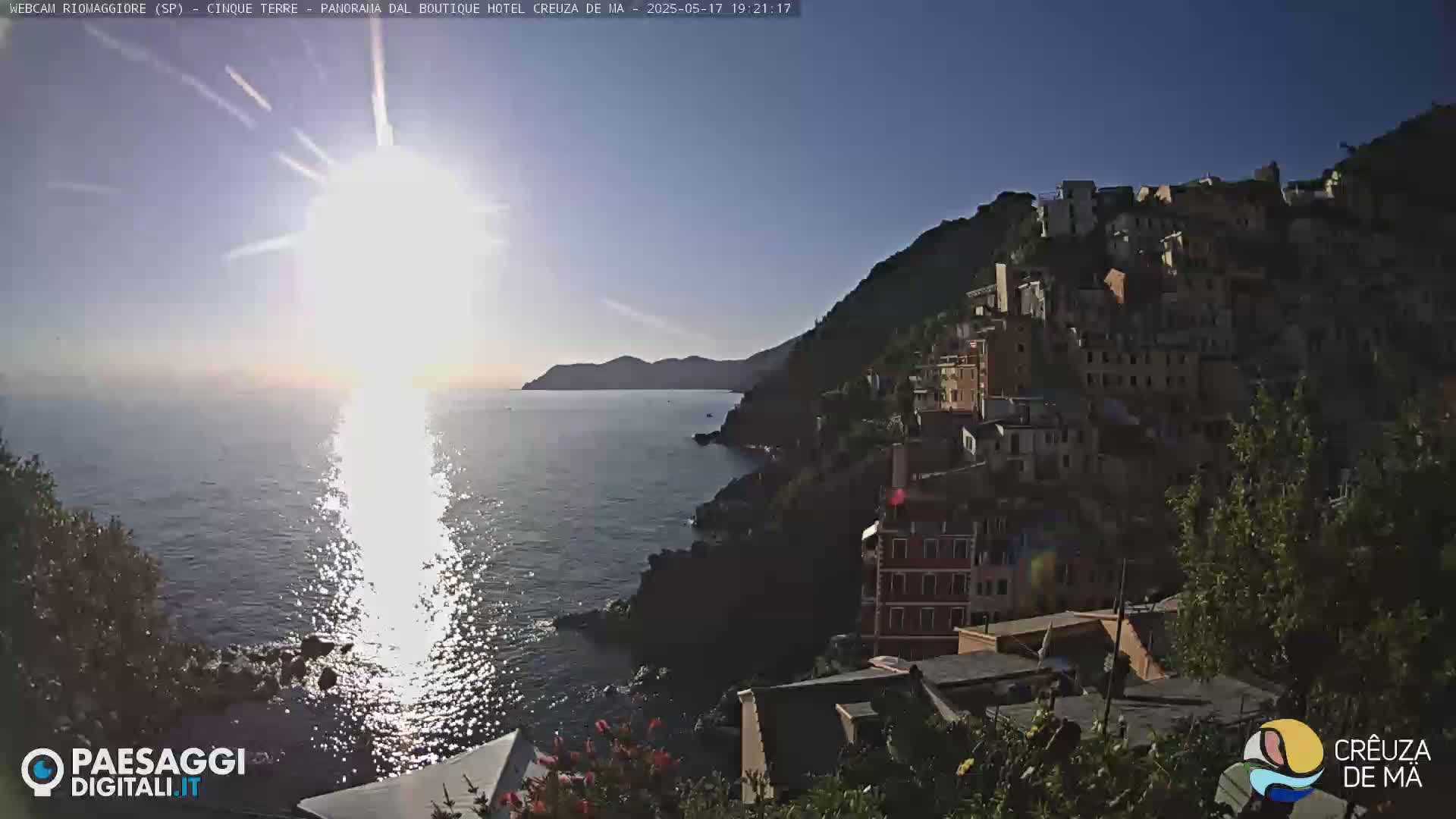 A bright sun shines over a calm ocean, reflecting its light on the water, with a hillside village of colorful buildings nestled into the cliffside.