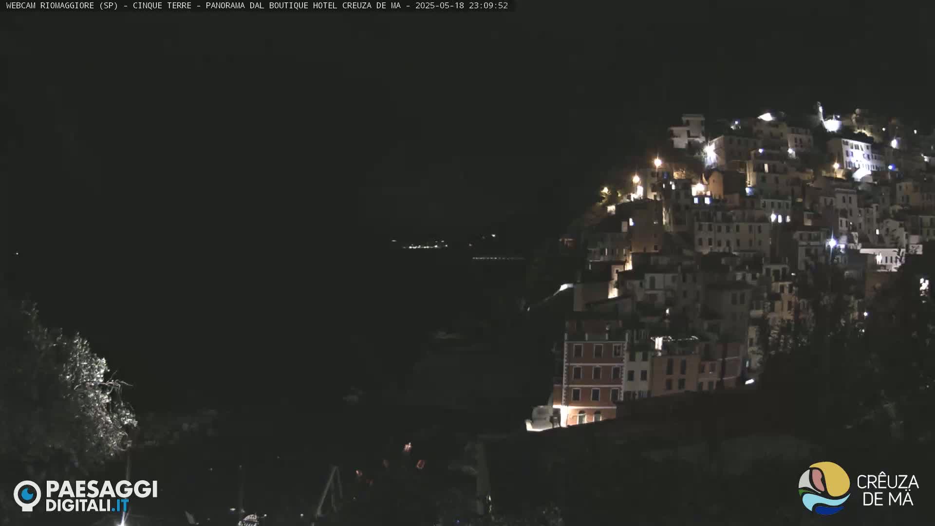 A nighttime view of a hillside town illuminated by lights, overlooking a dark body of water under a clear, dark sky.