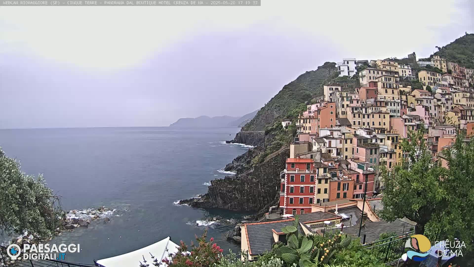 A coastal village of colorful buildings clings to a steep hillside overlooking a calm, gray sea under an overcast sky.