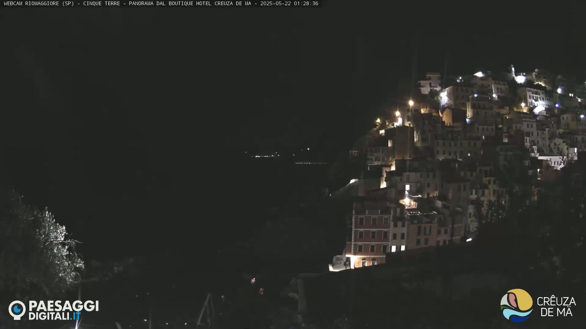 A nighttime view of a hillside town illuminated by lights, overlooking a dark body of water.
