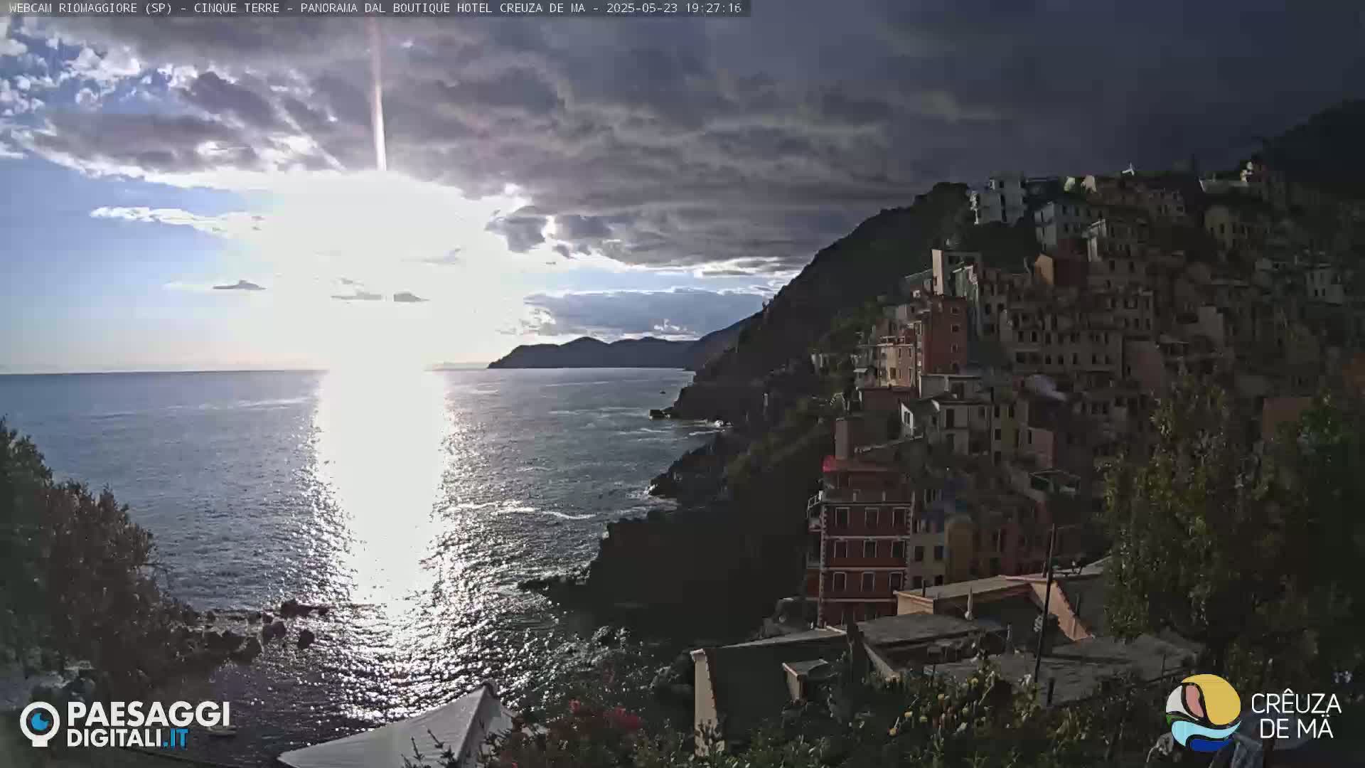 A coastal town clings to a steep cliffside overlooking a calm sea reflecting the setting sun under a partly cloudy sky.