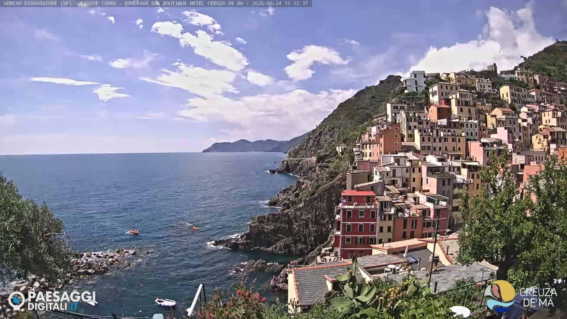 A picturesque coastal town, built on a steep hillside, overlooks a calm blue sea under a partly cloudy sky.