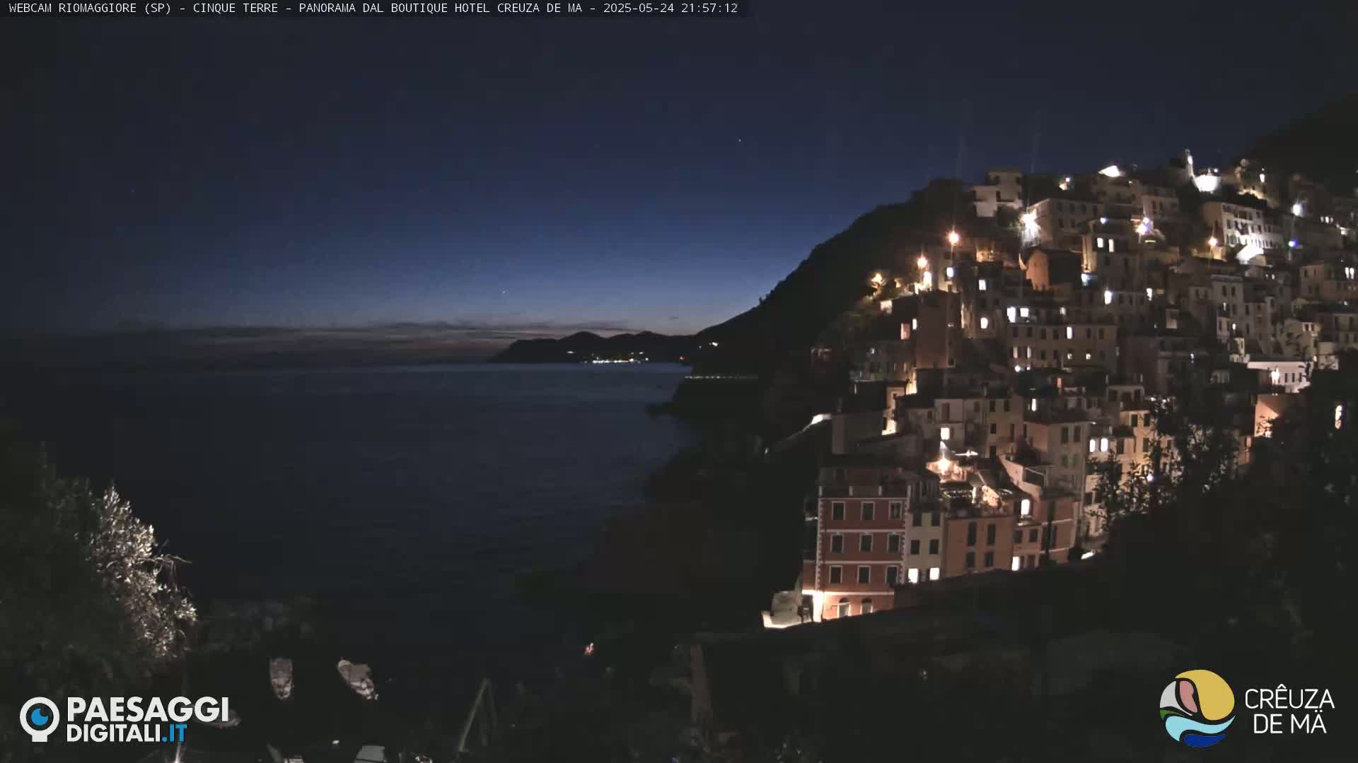 A nighttime view of a hilltop town illuminated by lights overlooking a dark sea under a twilight sky.