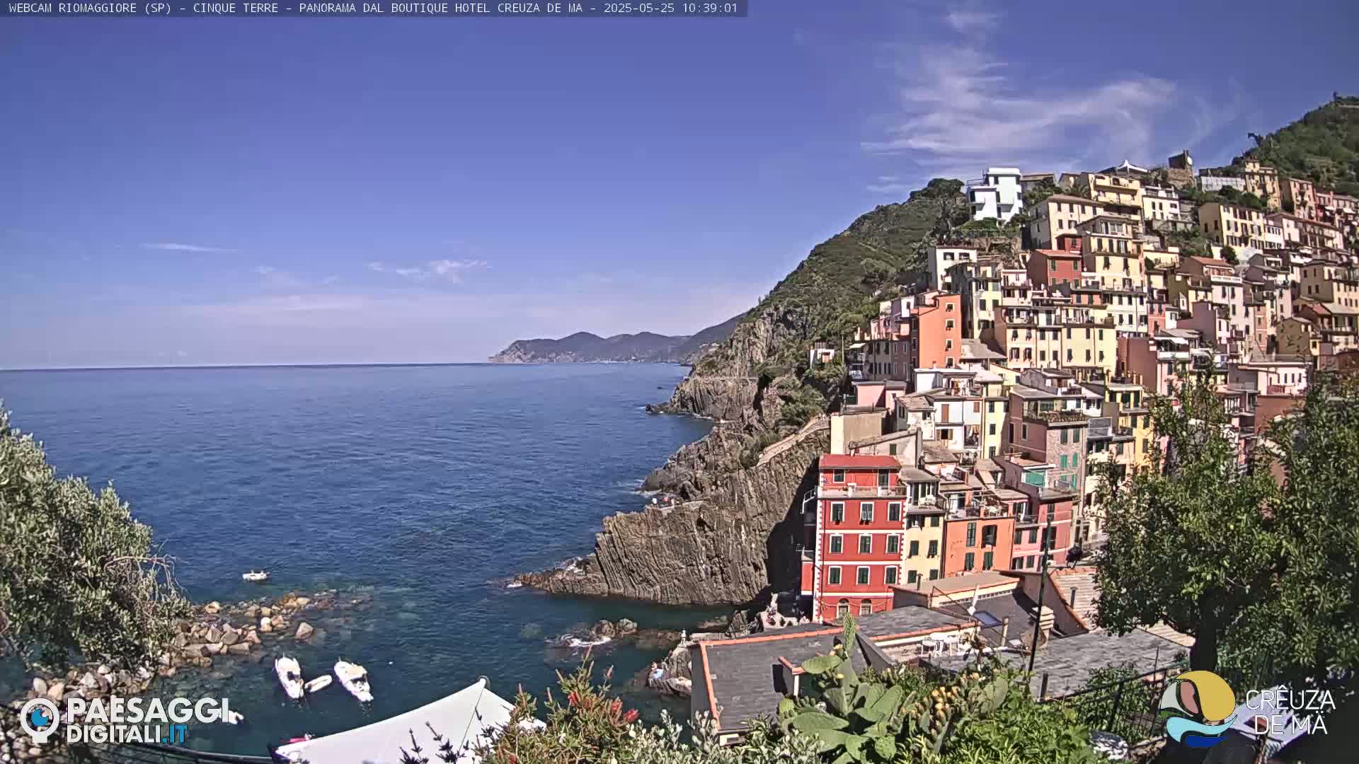 A picturesque coastal village clings to a hillside overlooking a calm, blue sea under a mostly clear sky.