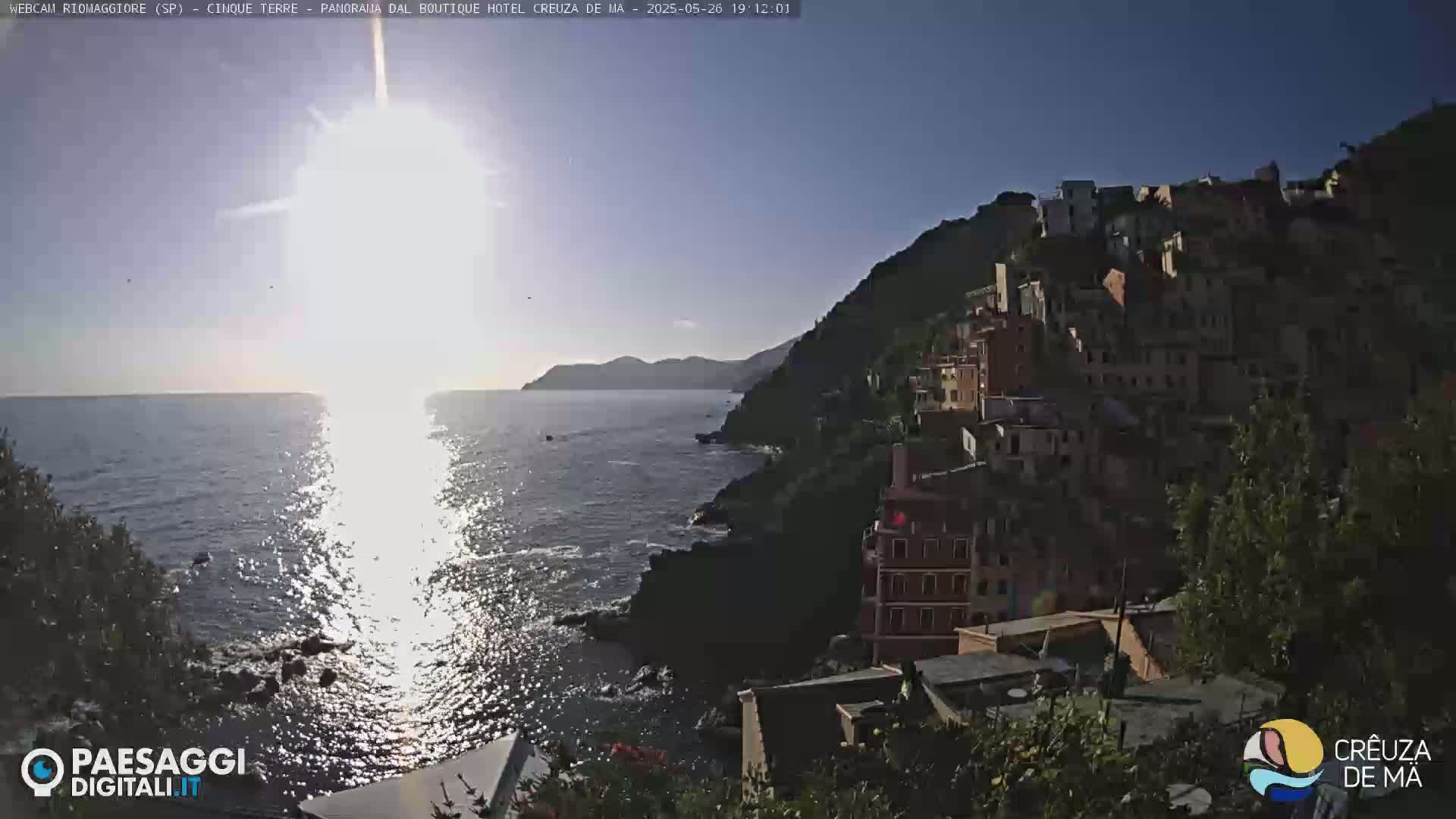 A sunlit coastal village clings to a hillside overlooking a sparkling sea under a clear blue sky.