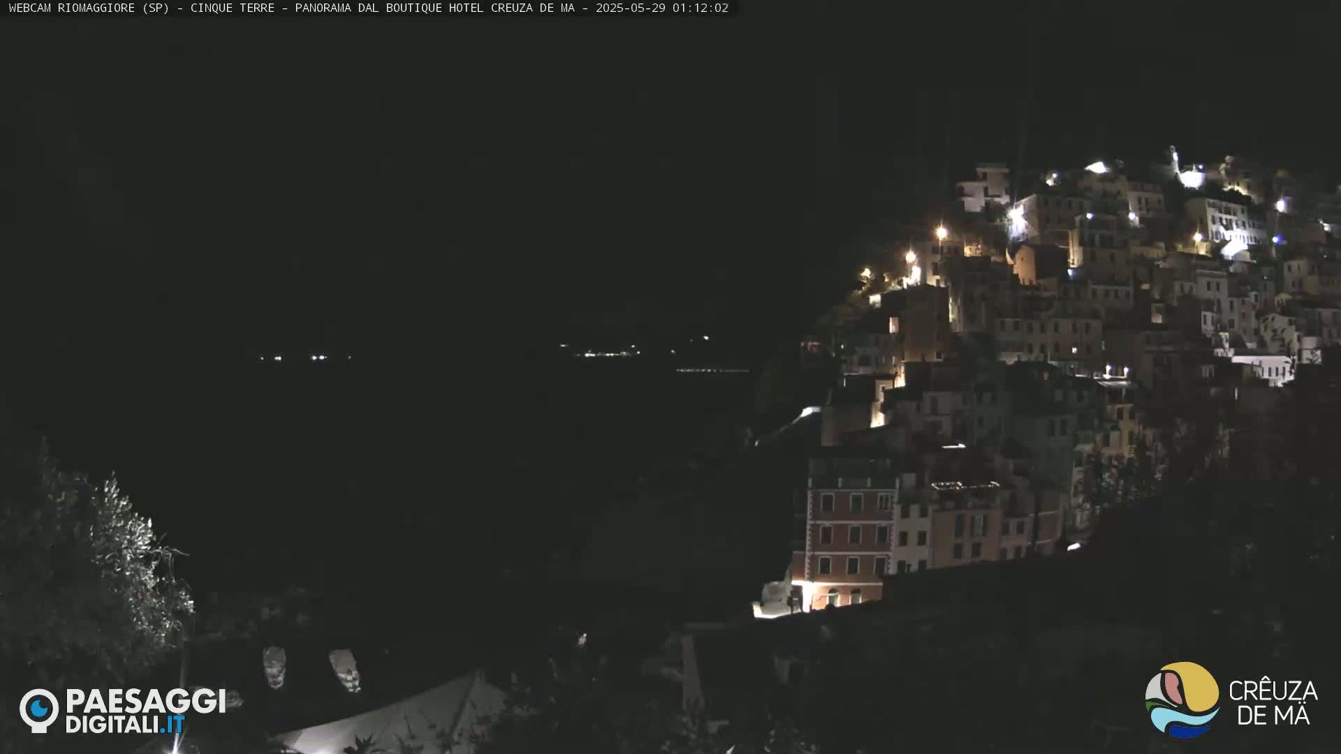 A nighttime view of a hillside town with many buildings lit up, overlooking a dark body of water under a clear, dark sky.