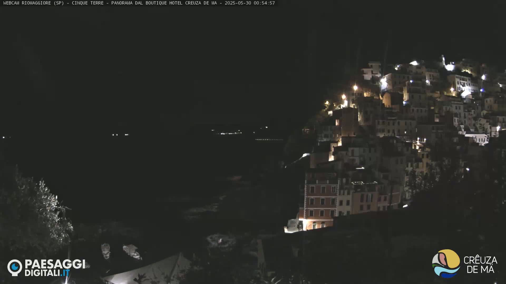 A nighttime view of a coastal town with many lights illuminating buildings on a hillside overlooking dark water.