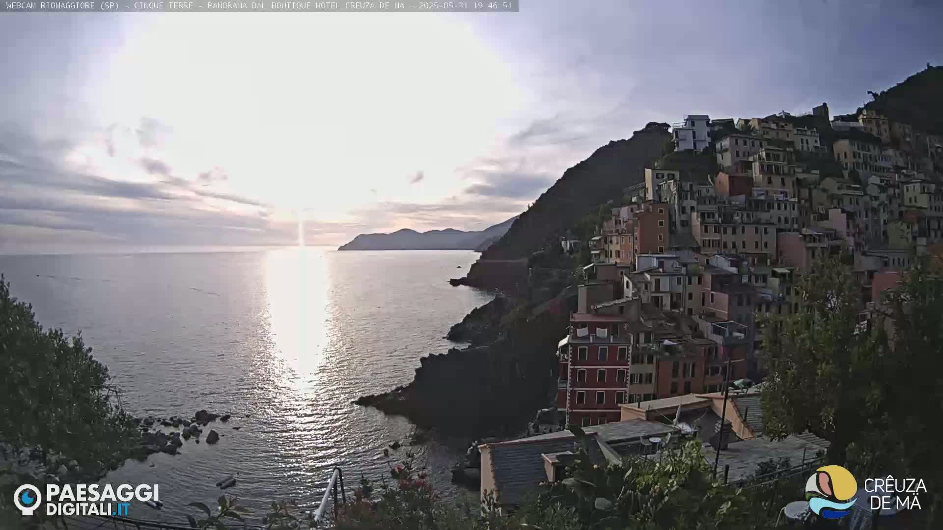 A coastal town built into a hillside overlooks a calm sea at sunset under a mostly clear sky.