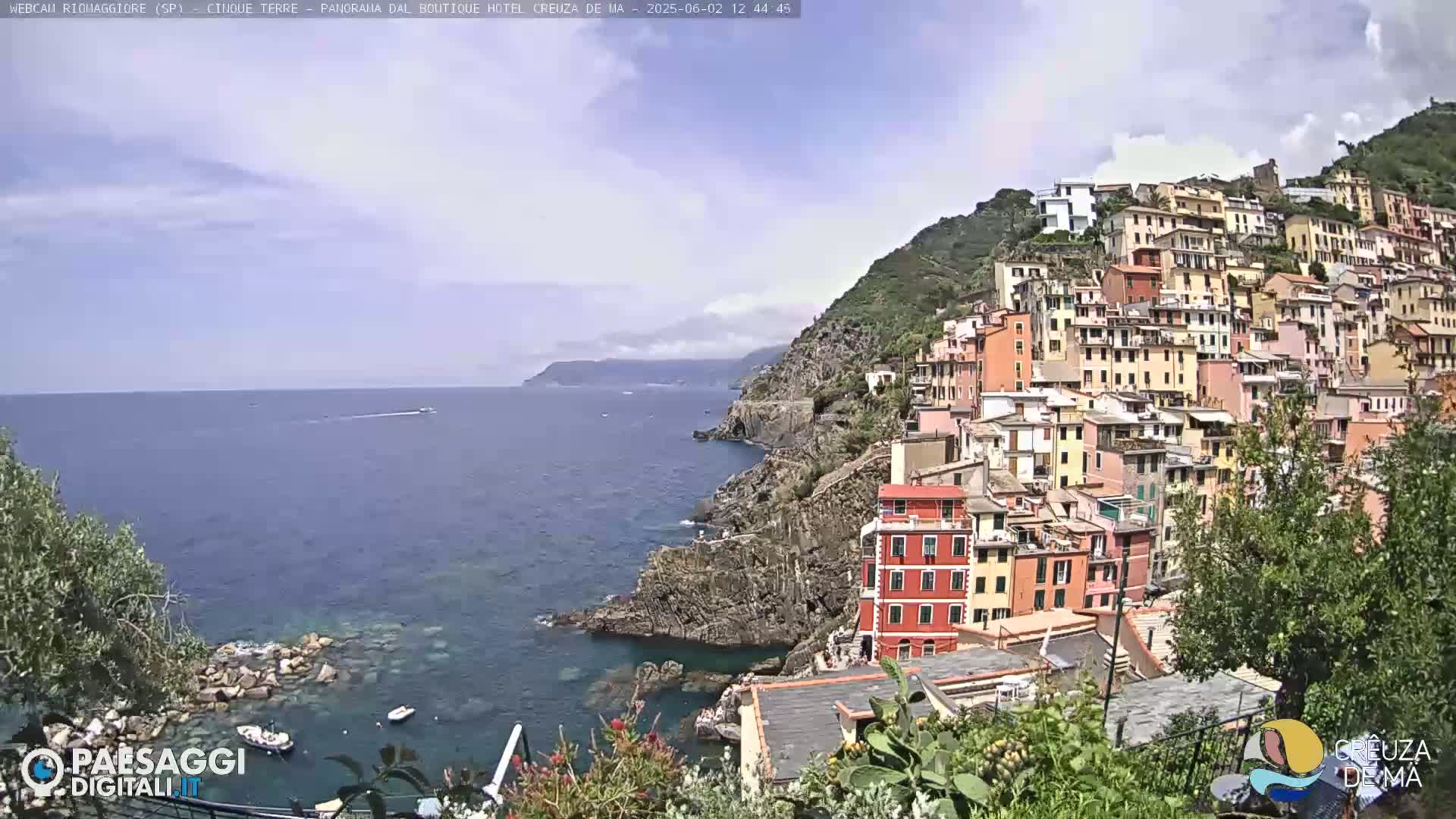 A picturesque coastal village of colorful buildings clings to a steep hillside overlooking a calm sea under a partly cloudy sky.