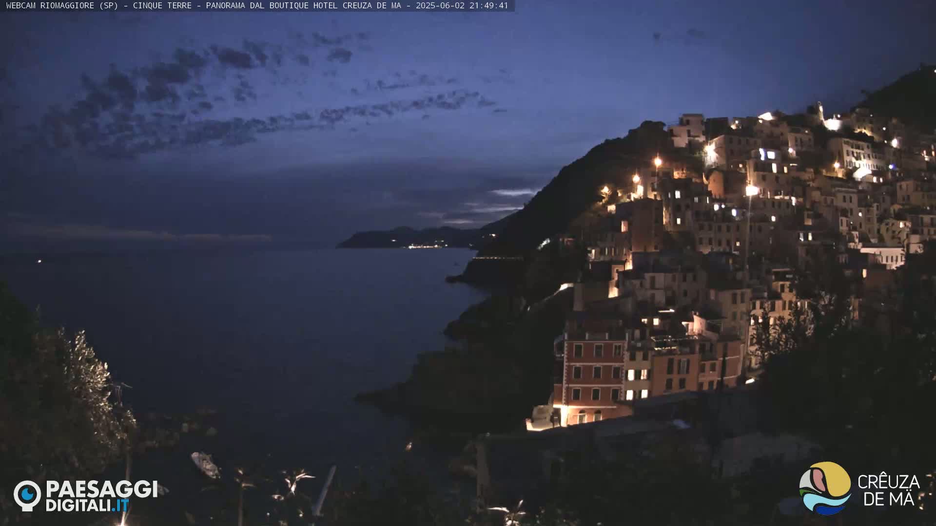 A hilltop town, illuminated at night, overlooks a dark, calm sea under a partly cloudy twilight sky.
