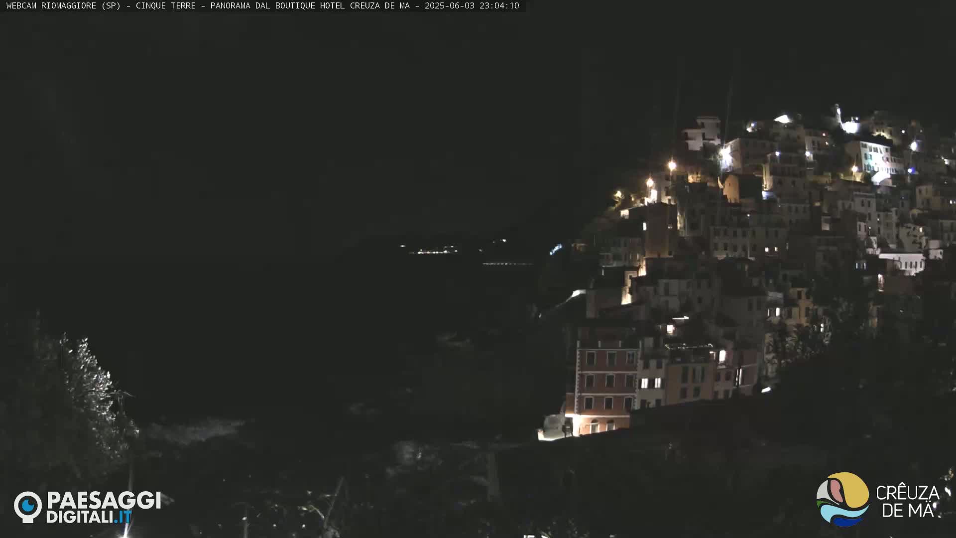 A nighttime view of a hillside town illuminated by lights, overlooking a dark body of water under a clear sky.