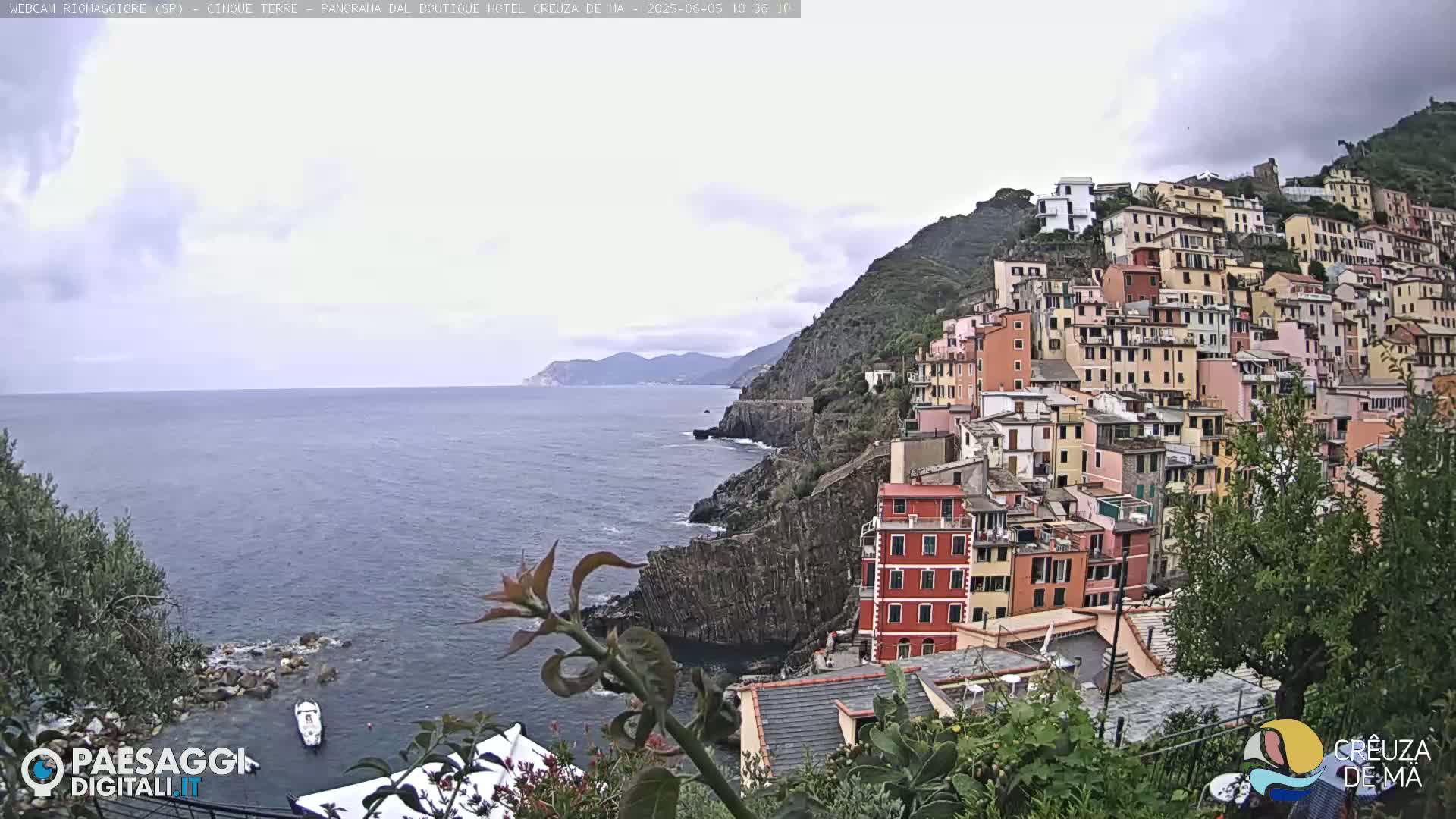 A coastal village of colorful buildings clings to a steep hillside overlooking a calm sea under an overcast sky.