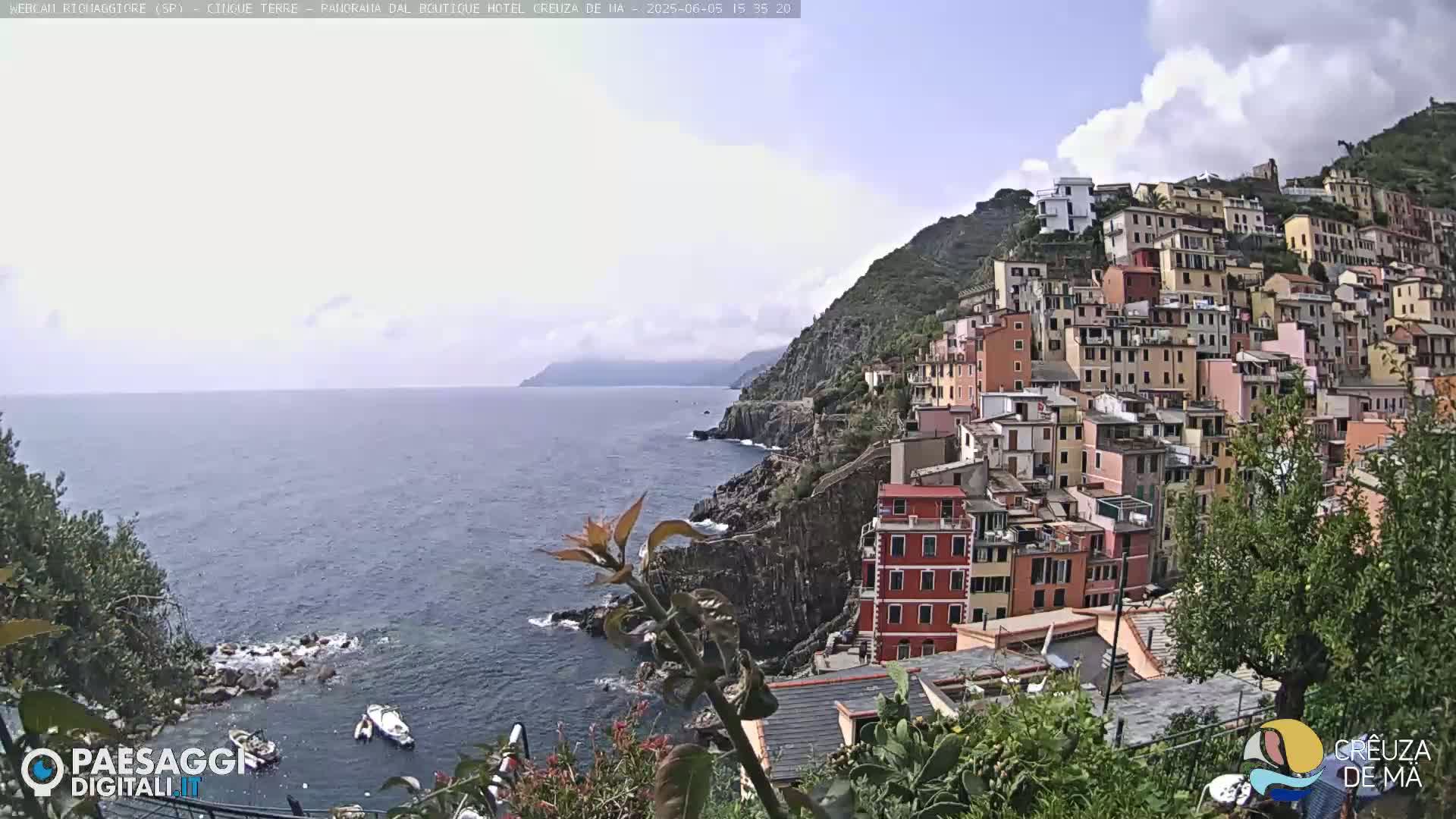 A coastal village of colorful buildings climbs a steep hillside overlooking a calm sea under a partly cloudy sky.