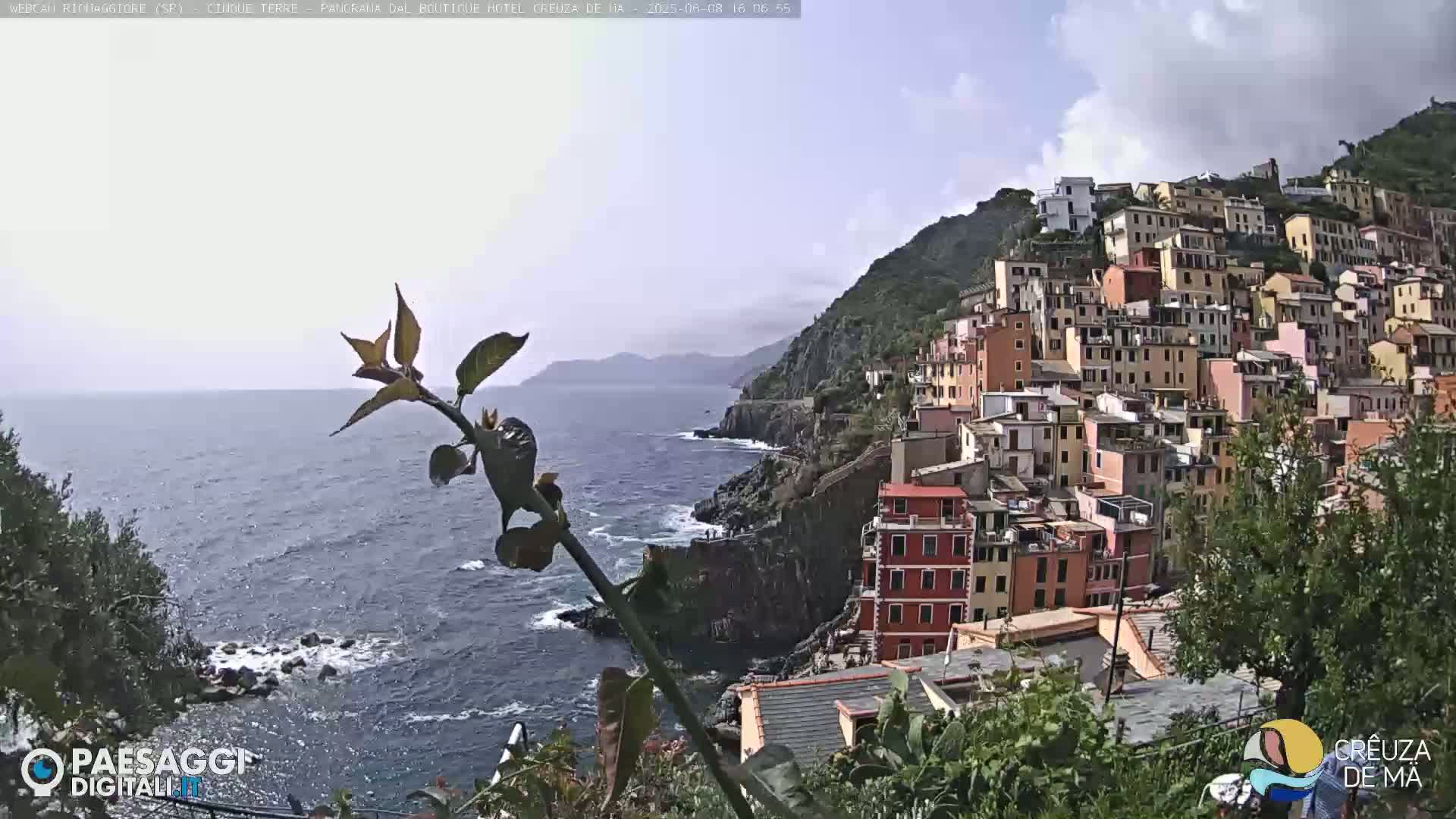 A coastal town with colorful buildings clinging to a hillside overlooks a blue sea under a partly cloudy sky.